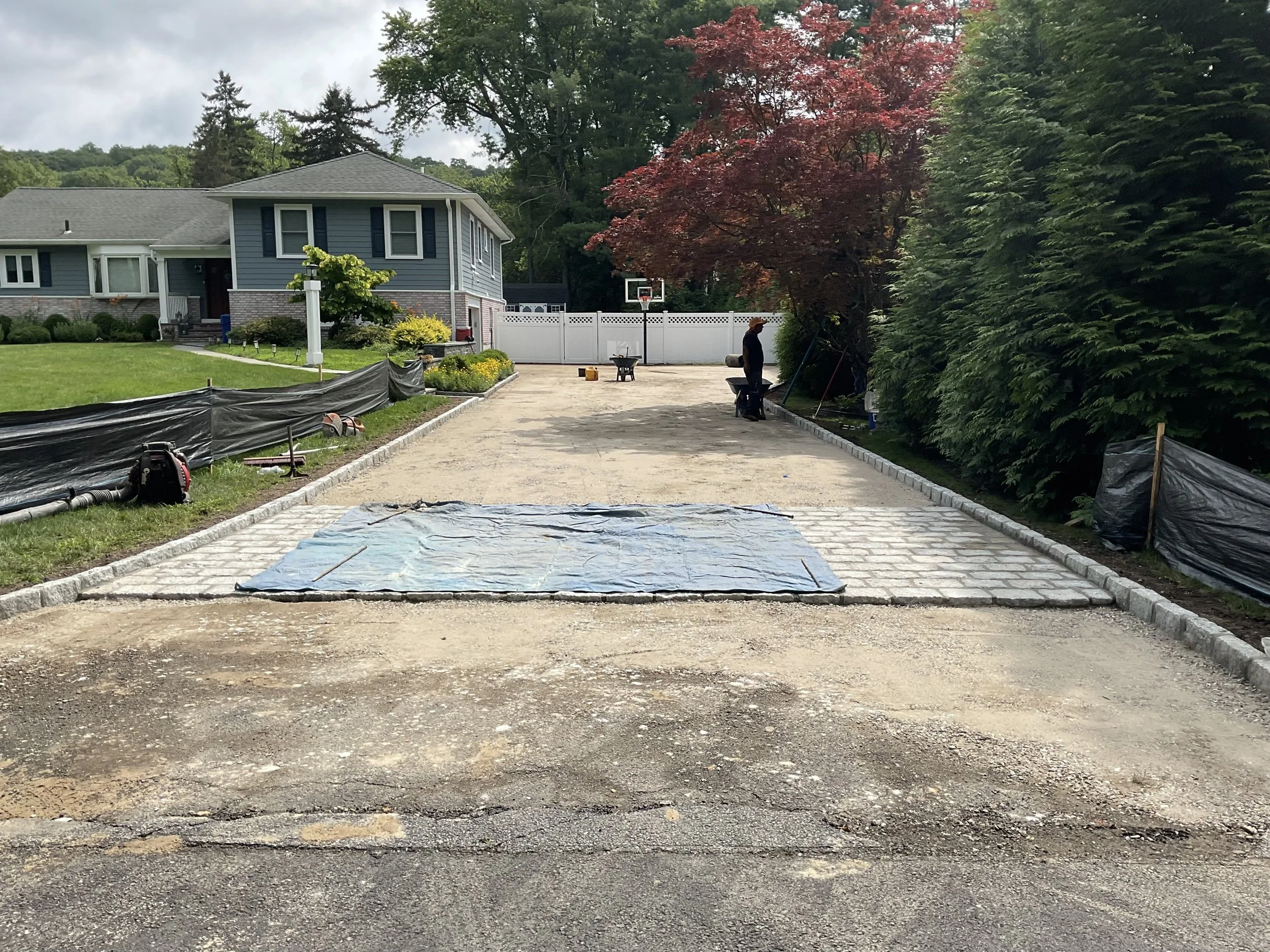 Under construction driveway with partially laid bricks, surrounded by green lawn, trees, and a house in the background.