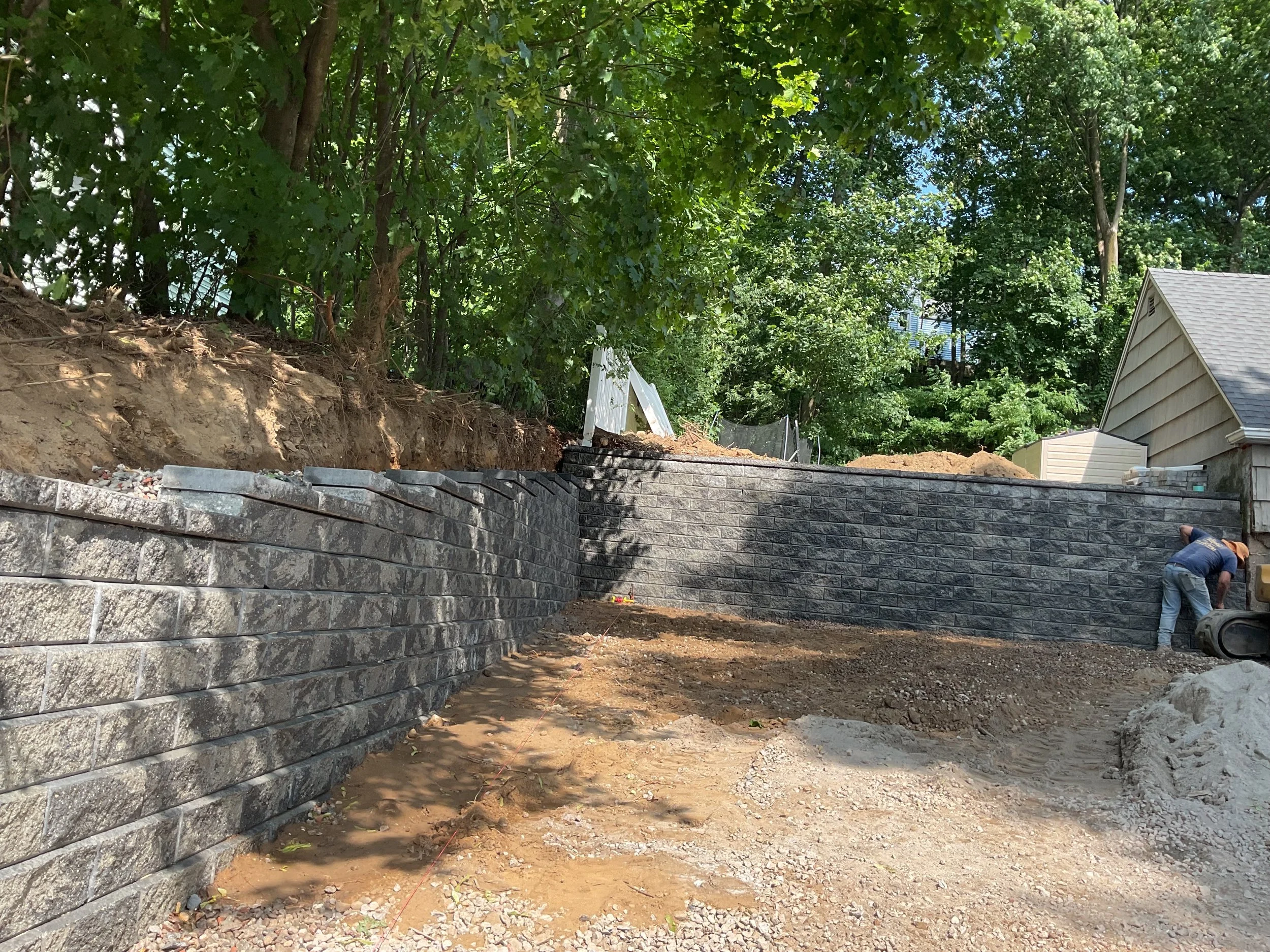Construction workers building a stone retaining wall in a yard with dirt and trees surrounding the area.