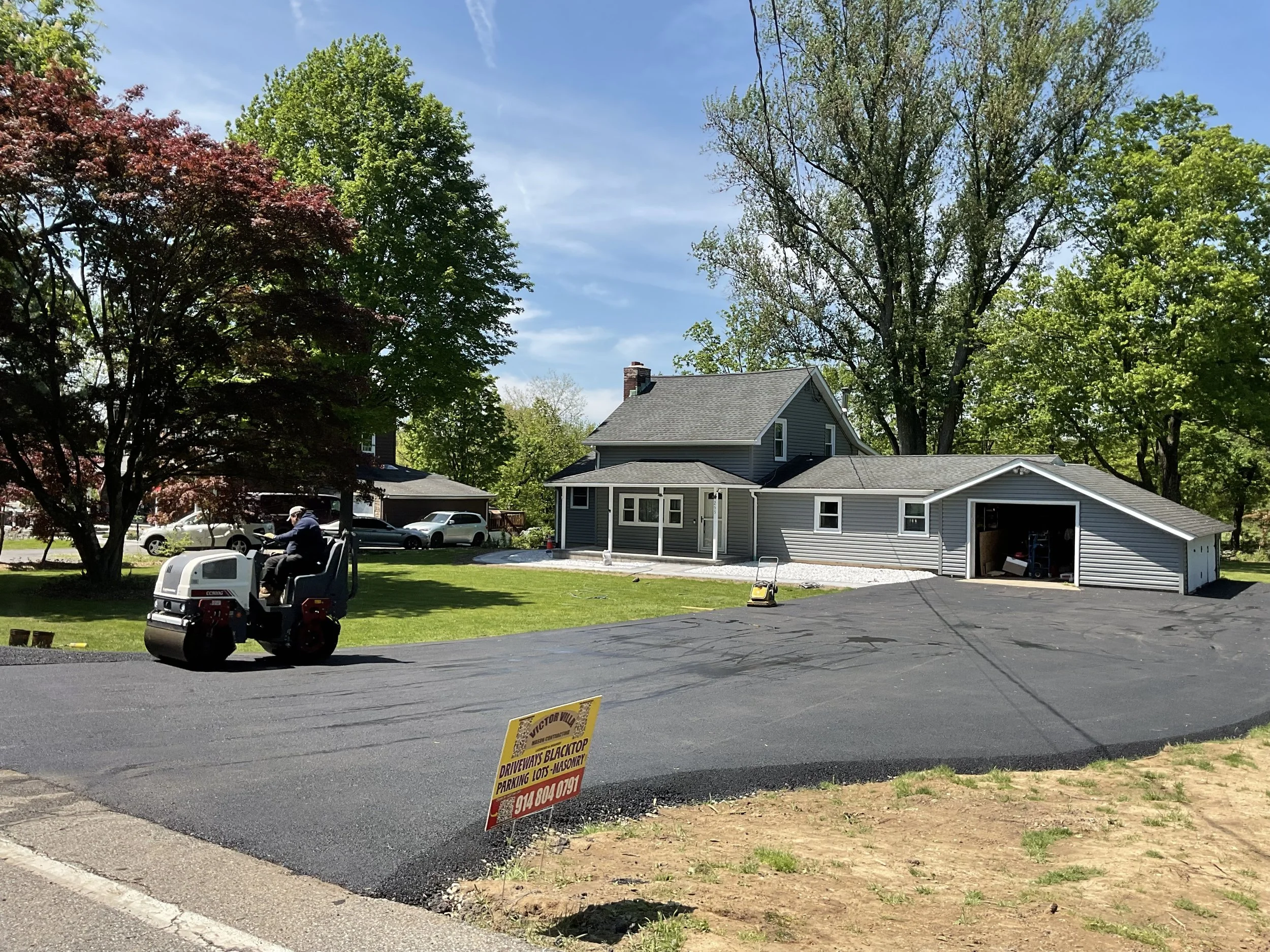A homeowner installing a new blacktop driveway with a compact roller machine, a house with grey siding, a garage, surrounded by green trees, and a sign for driveway, parking lot, and masonry services.