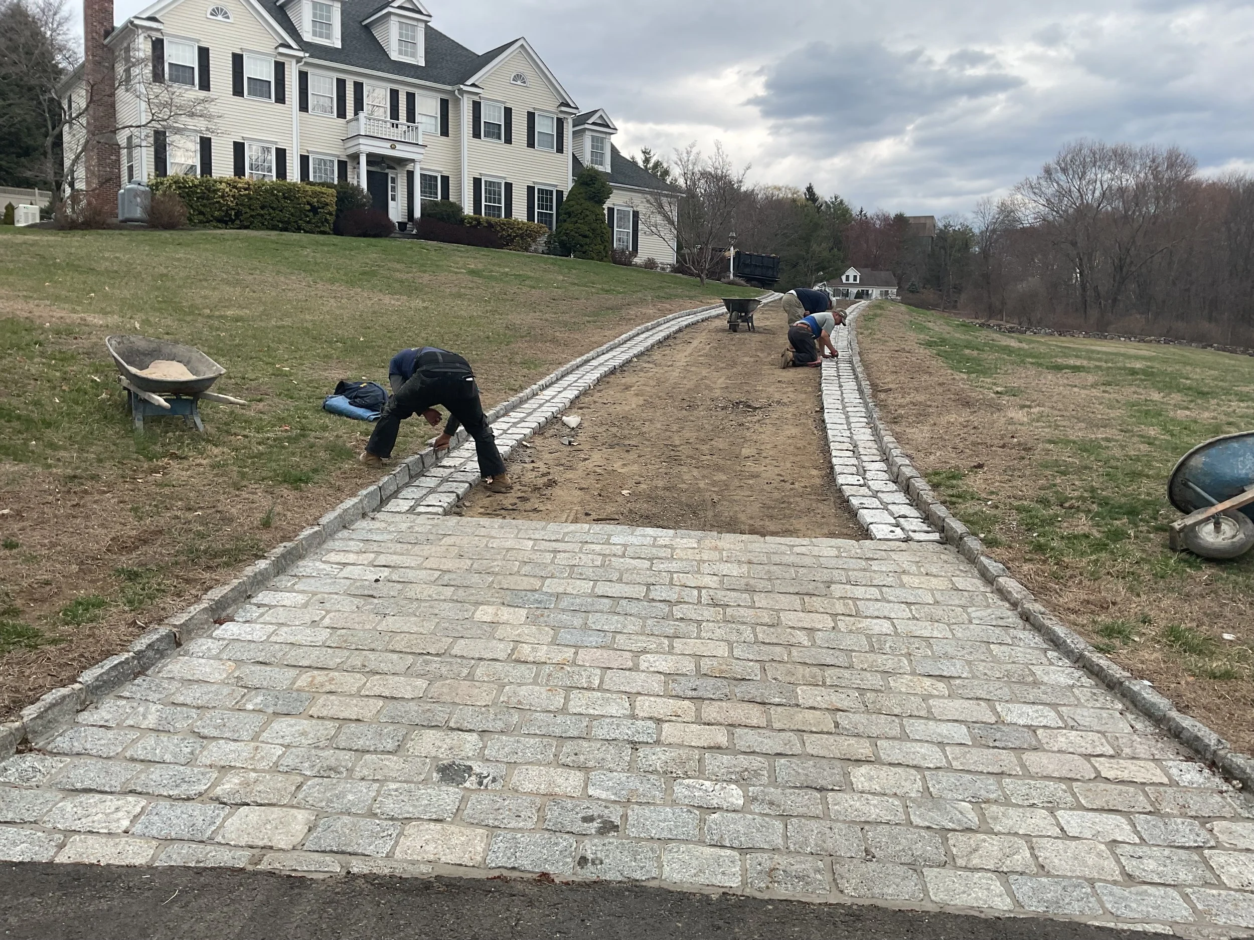 Workers installing a brick pathway leading up a hill in front of a large white house with black shutters. The pathway is in the process of being laid, with workers placing bricks along the edges. The scene shows a cloudy sky and a grassy area on eith