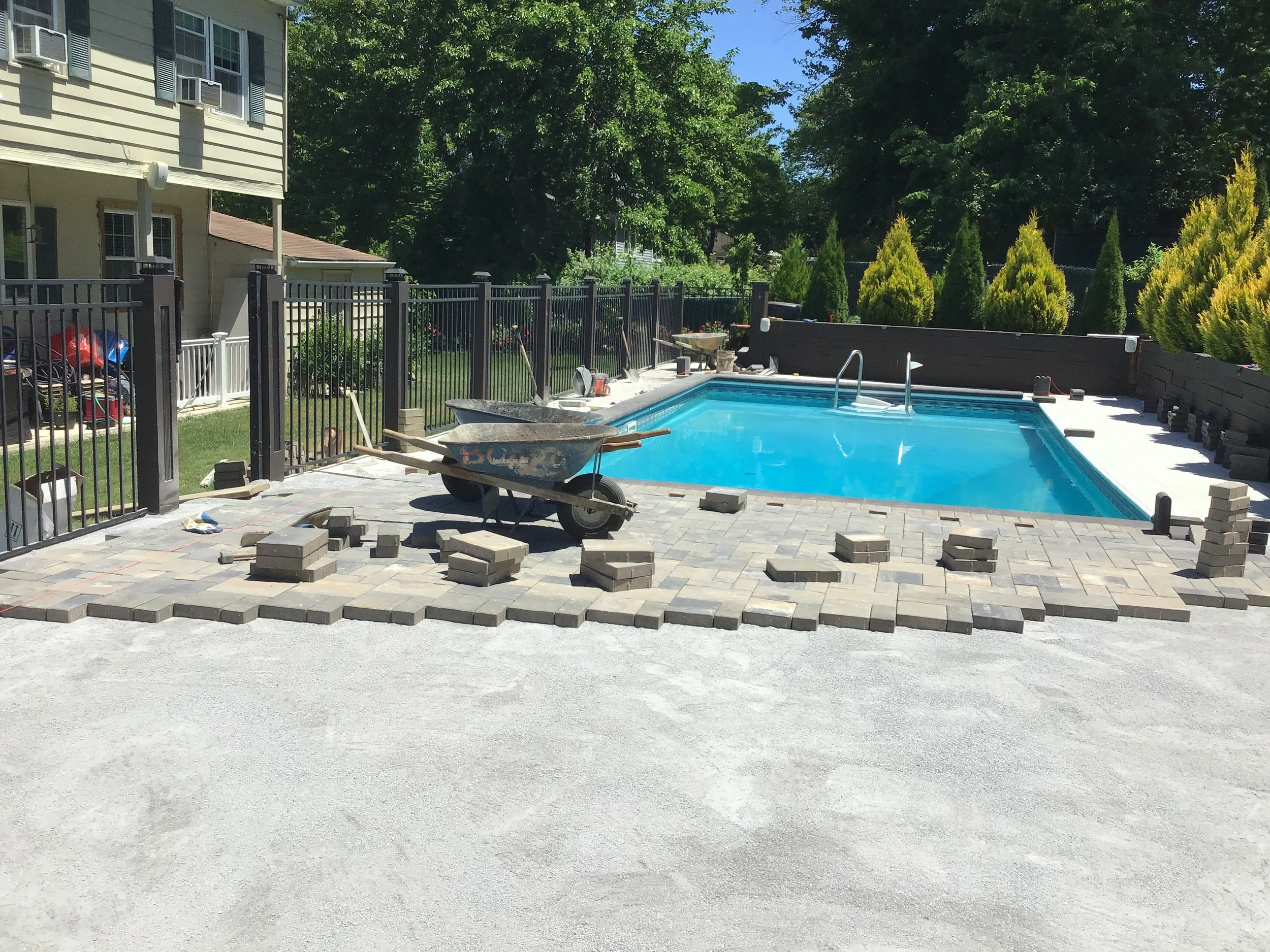 In-ground swimming pool under construction with paving stones being laid, a wheelbarrow, and stacks of bricks nearby, surrounded by a black metal fence and a landscape with yellow-green bushes and trees.