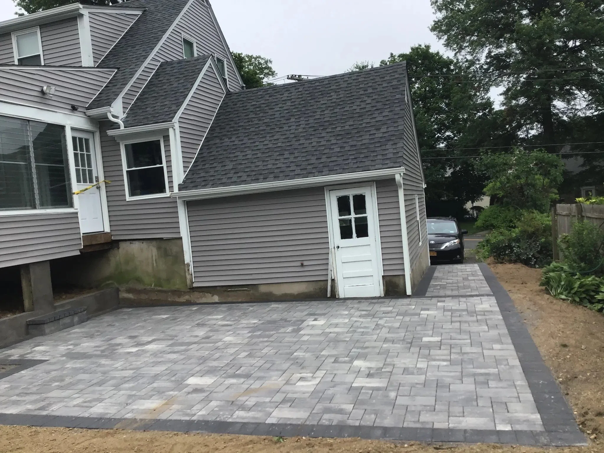 Residential backyard with new gray brick driveway and a small gray shed with a white door, adjacent to a multi-story house with gray shingles, with a parked car in the background and trees.
