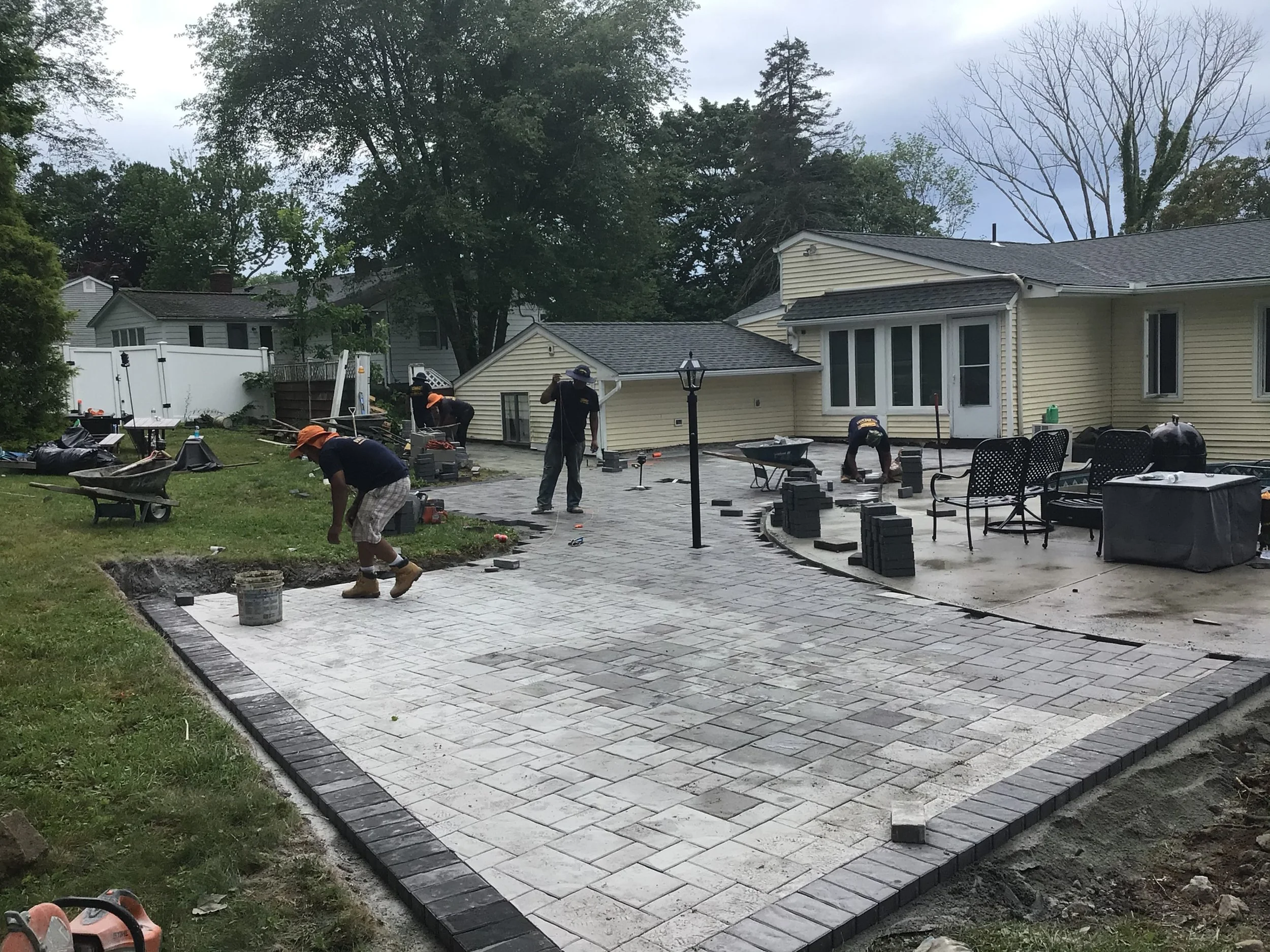 Workers are laying pavers for a backyard patio with stacks of bricks, wheelbarrows, and outdoor furniture visible.