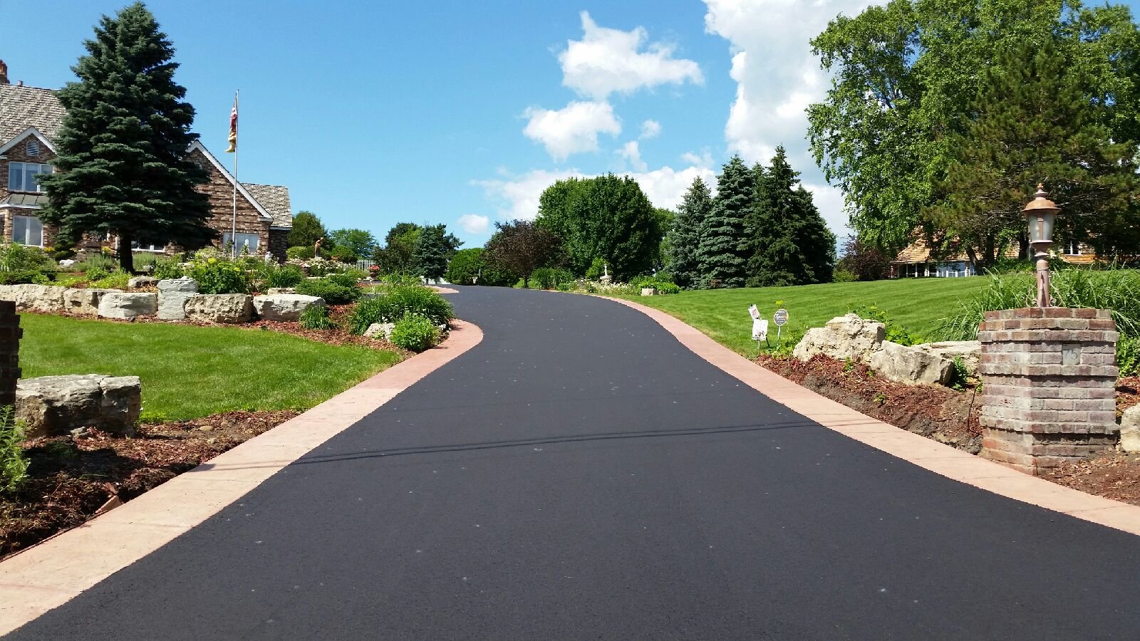 A winding paved driveway bordered by a brick sidewalk, with landscaped gardens, large rocks, green grass, tall trees, and a house with a flag in the background on a sunny day with a partly cloudy sky.