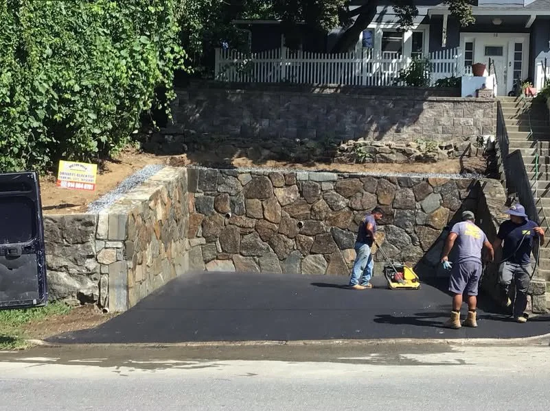 Construction workers are paving asphalt on a driveway in front of a stone retaining wall, with a house and stairs visible in the background.