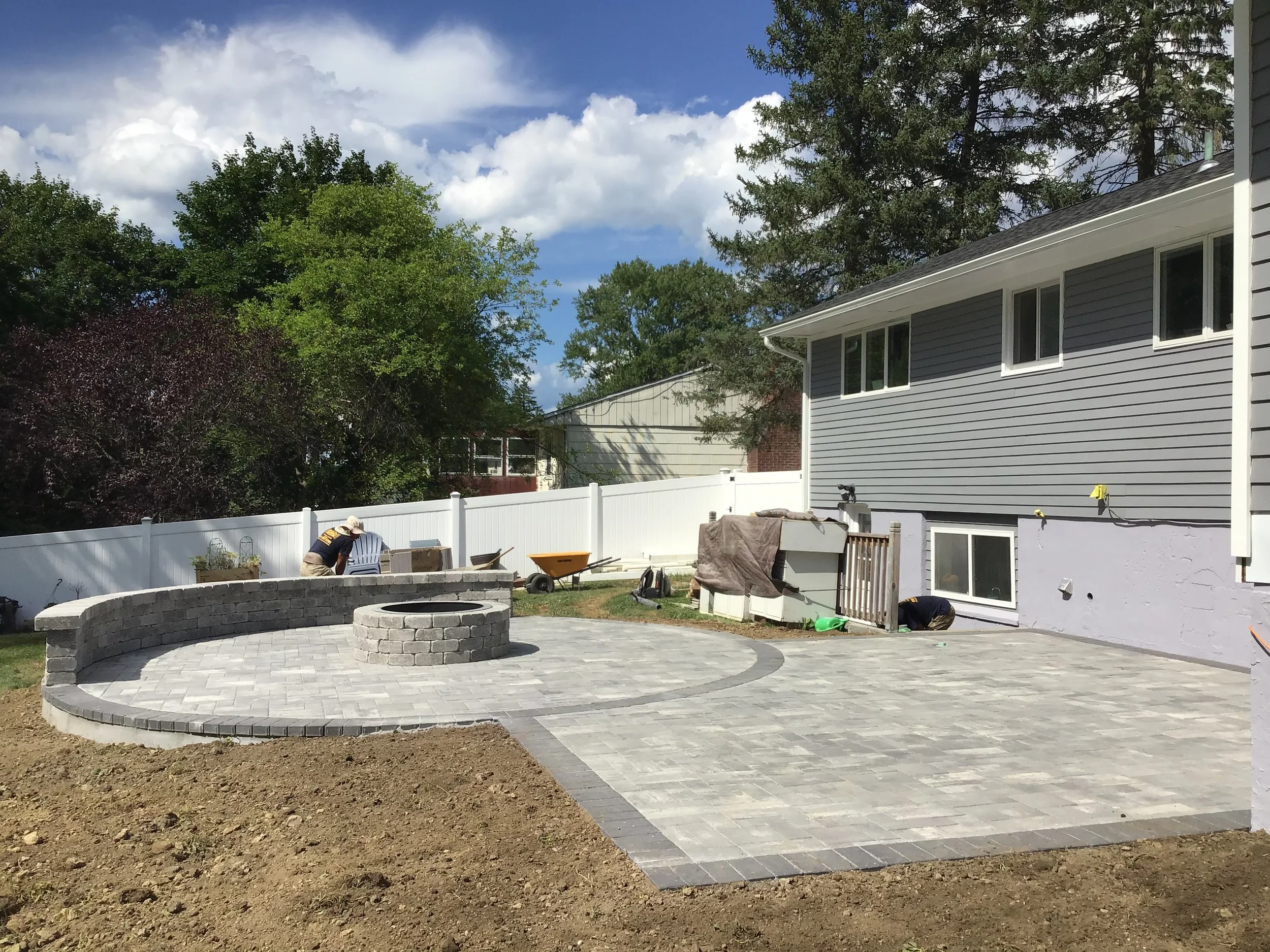 Construction of a new backyard patio with a circular firepit area, gray interlocking pavers, a white privacy fence, and a gray house with white trim. Workers are installing the pavers and working near the house.