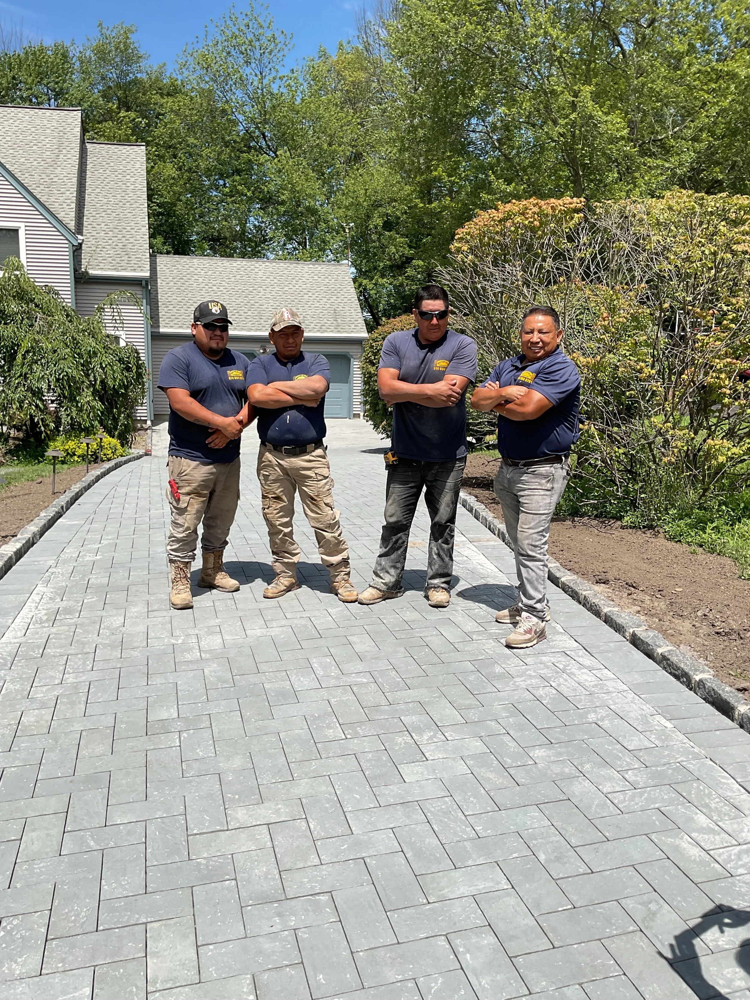 Four men standing with arms crossed on a paved walkway outdoors, with trees, bushes, a house, and a blue sky in the background.