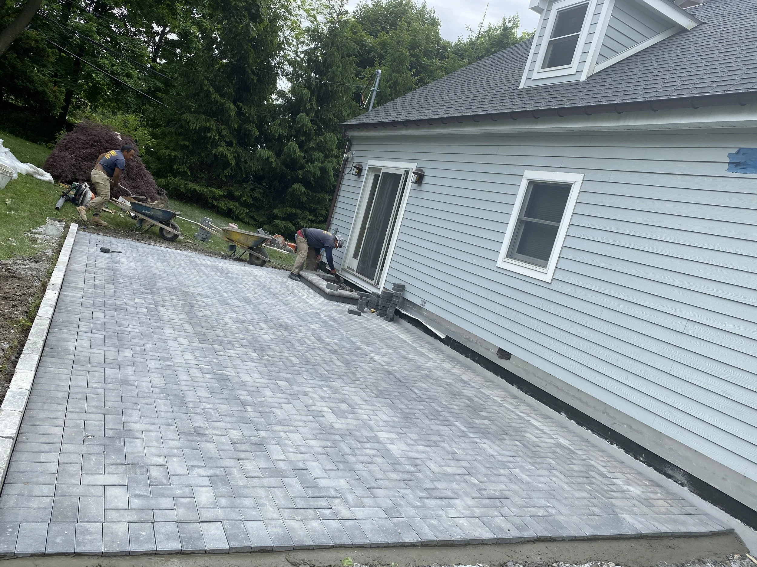Workers installing brick patio in backyard next to light blue house with a sliding glass door and two windows, surrounded by trees.