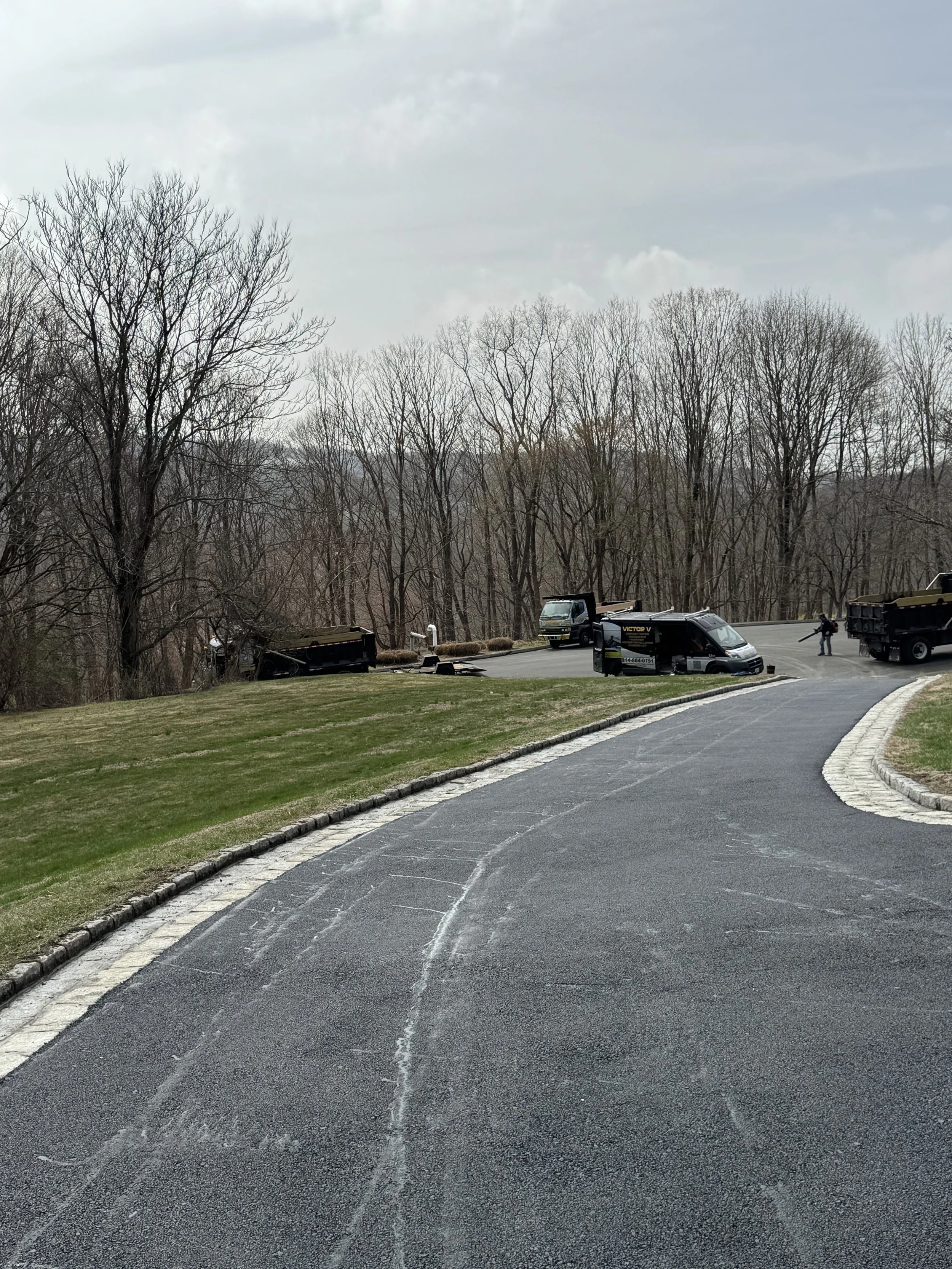 Several trucks and a person in a parking lot surrounded by leafless trees, with a winding paved driveway in the foreground.