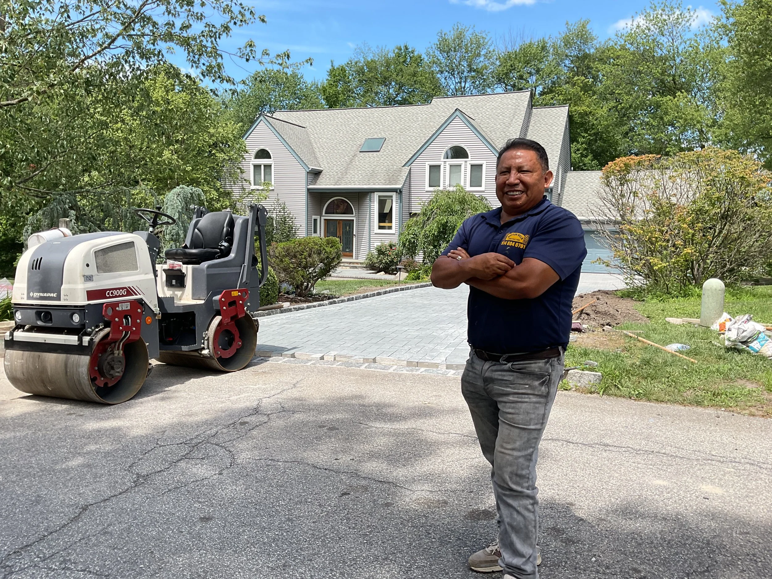A man stands outdoors in front of a residential house with a driveway, smiling and crossing his arms. A large steamroller vehicle is parked nearby, and lush trees and bushes surround the house.