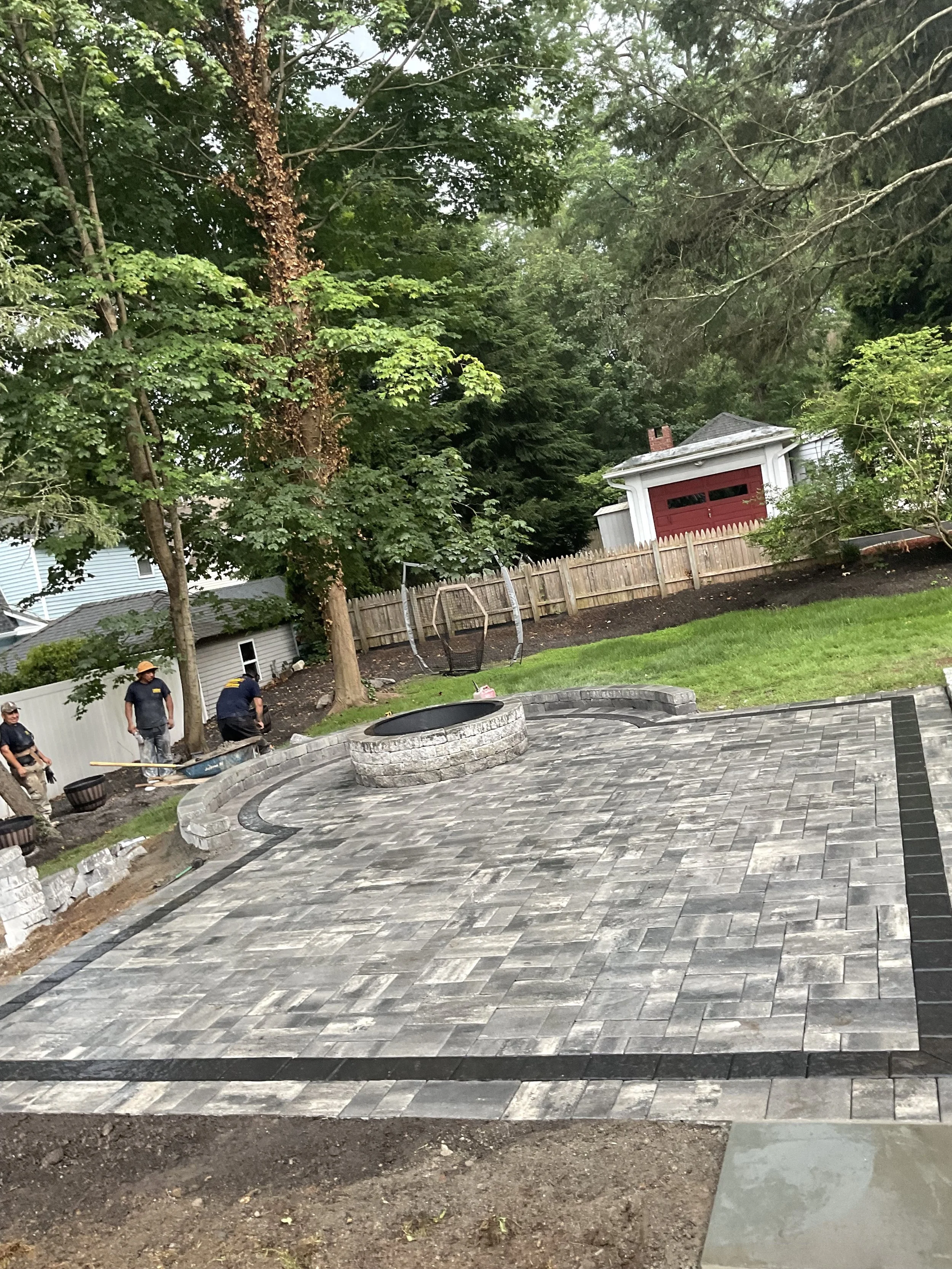 Backyard with newly laid stone patio, fire pit in center, and workers installing features. Green trees and a red garage in the background.