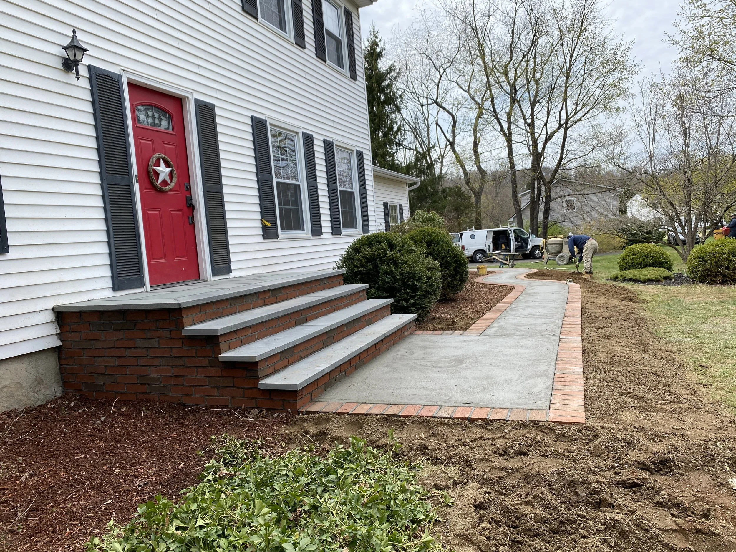 Front porch with red door, new concrete walkway with brick border, and workers planting garden beds in a residential yard.