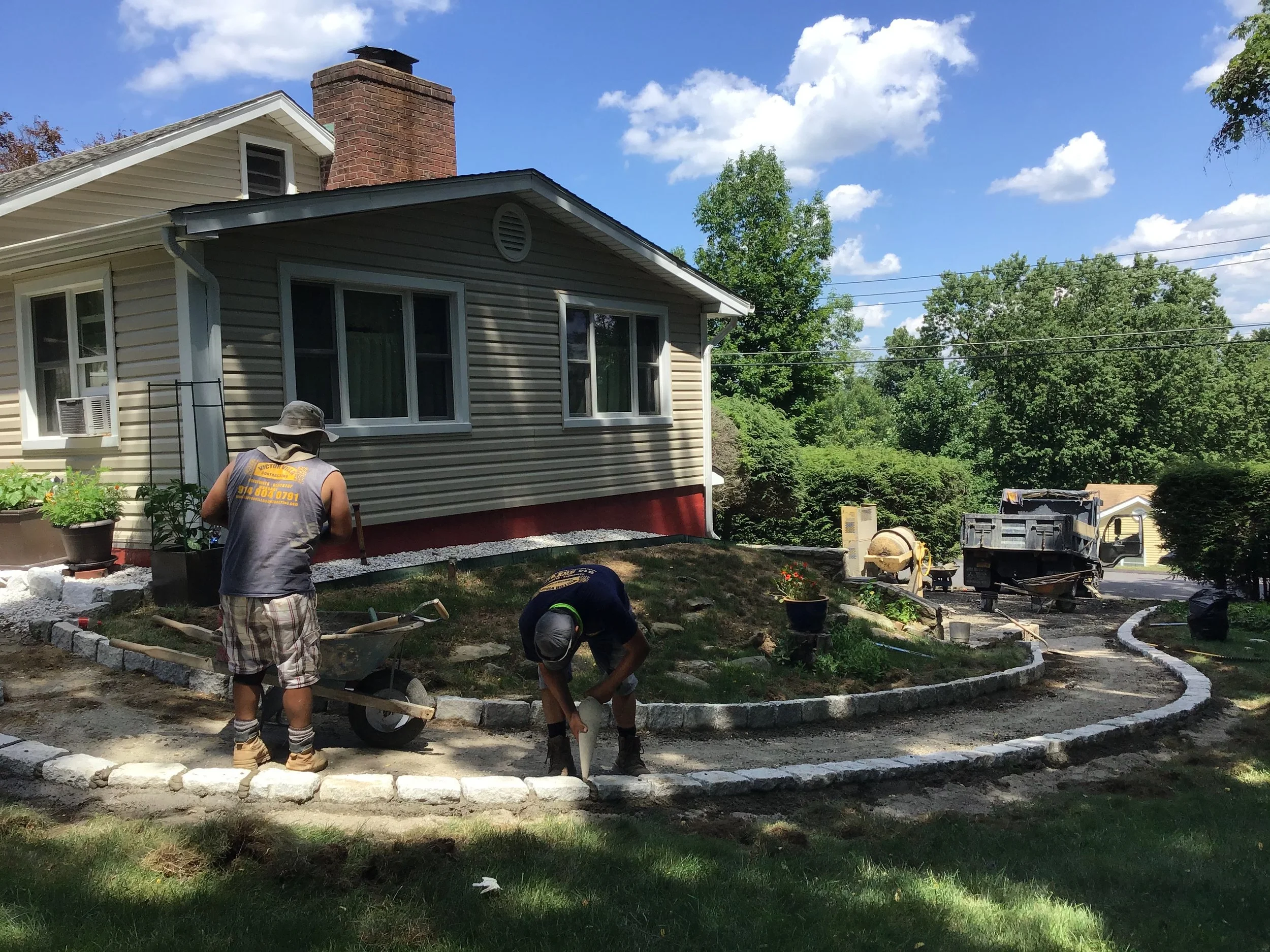 Two workers installing a stone pathway around a house on a sunny day. One is bending down, and the other is standing with a wheelbarrow nearby. The house is beige with white trim, and there are green trees in the background.