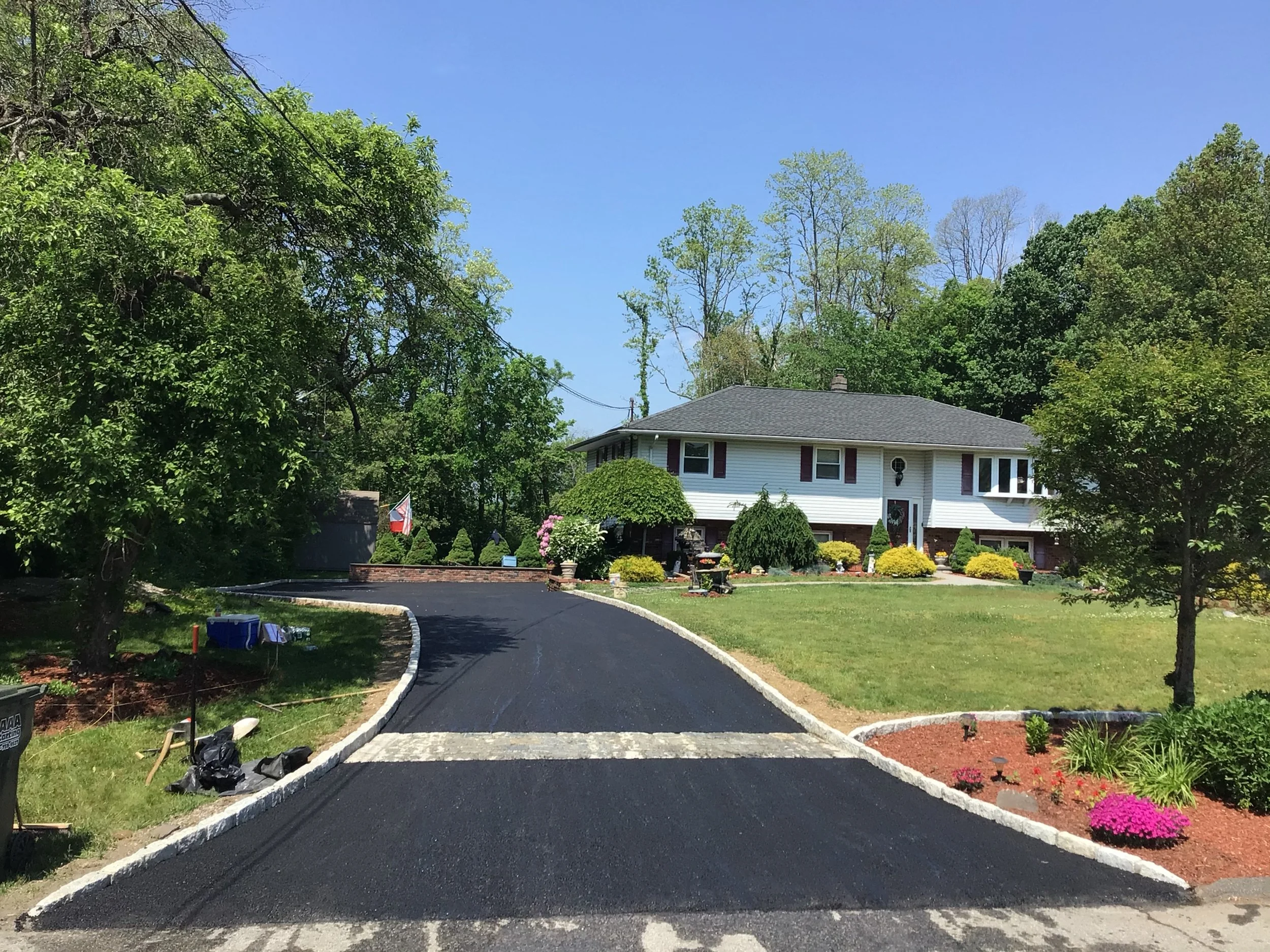 A newly paved black asphalt driveway leading to a white house with a well-maintained front yard decorated with colorful flowers and bushes. Tall green trees surround the house under a clear blue sky.