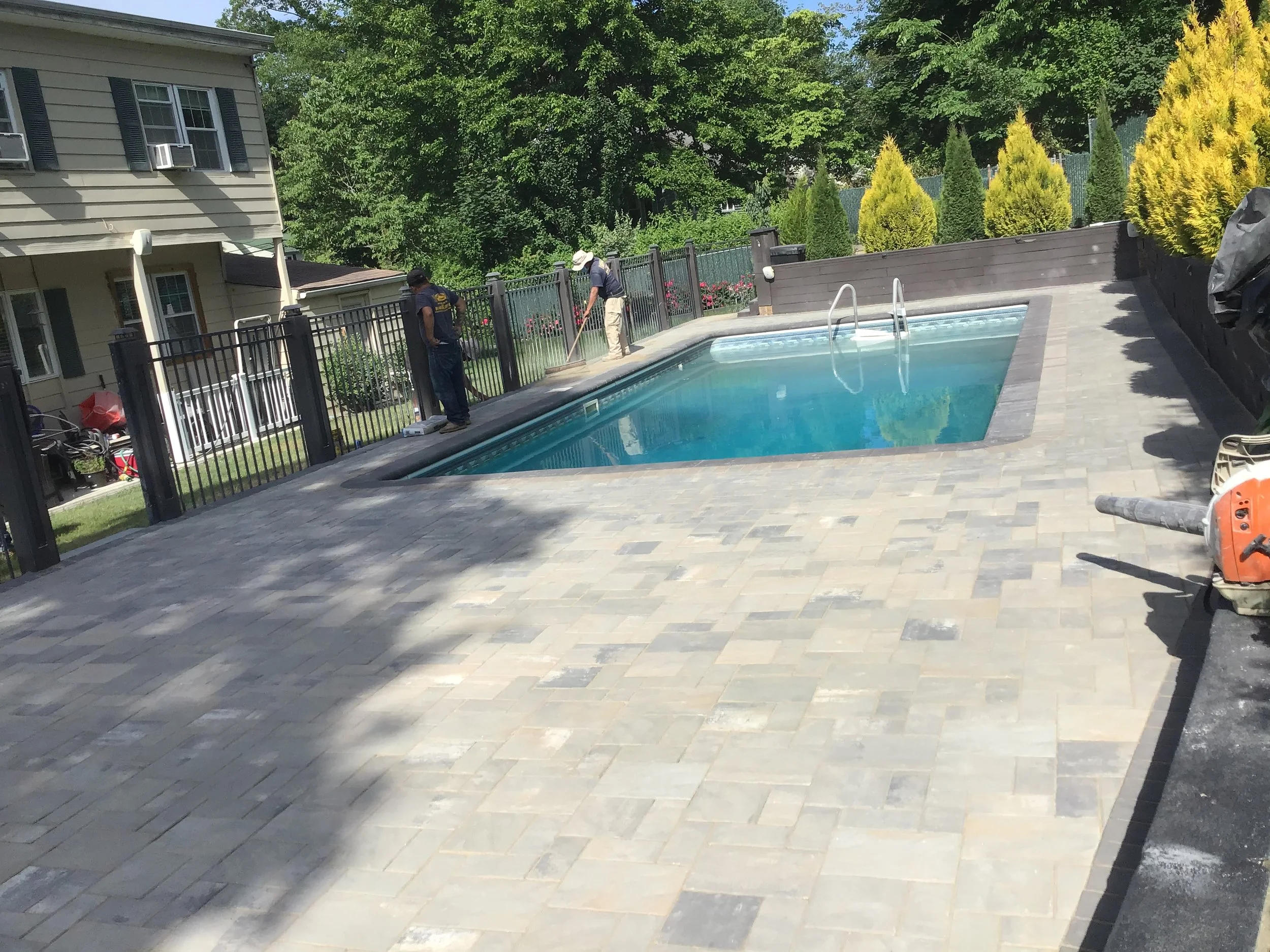 Workers installing a new paver patio around a backyard swimming pool, with a house and trees in the background.