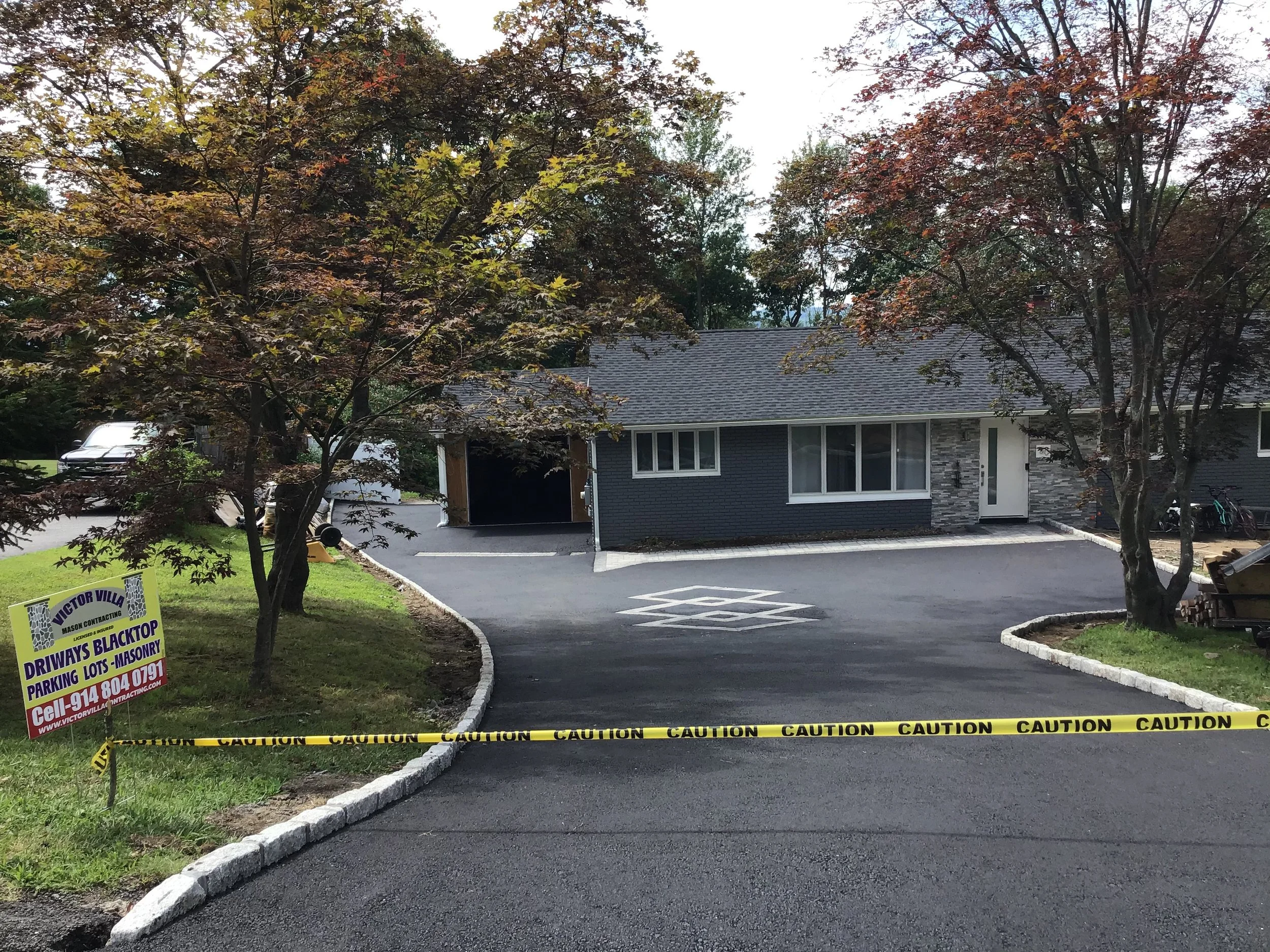 A newly paved driveway with caution tape, trees with red and green leaves, and a house in the background under cloudy sky, with a sign for Victor Villa Masonry and Drivways Blacktop.