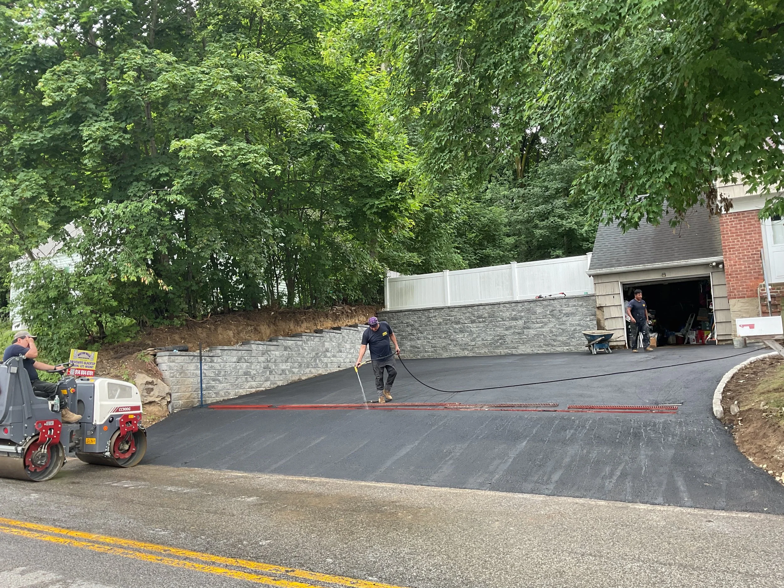 Workers paving a sloped driveway in front of a garage with a stone retaining wall and surrounded by trees.