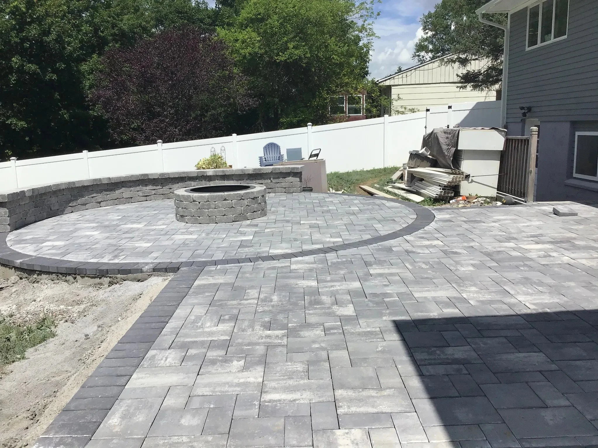 Backyard with recently paved stone patio, a circular fire pit, and a white fence surrounding the area. There are some stacked materials and a covered grill near the house, with trees and other houses in the background.