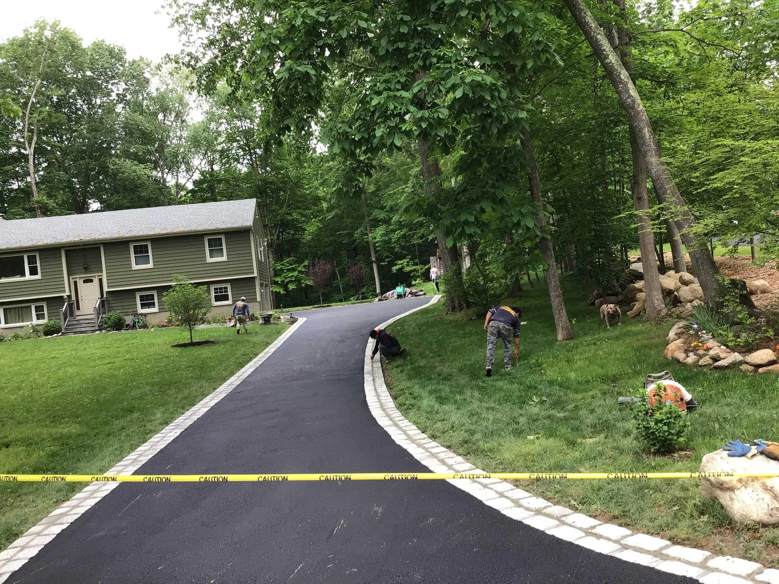 People are installing new grass on a lawn next to a freshly paved driveway in front of a green house, with trees and rocks in the background.