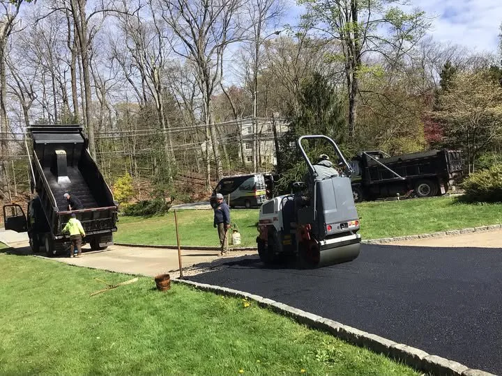 Workers paving a road with asphalt, surrounded by trees and a grassy area.