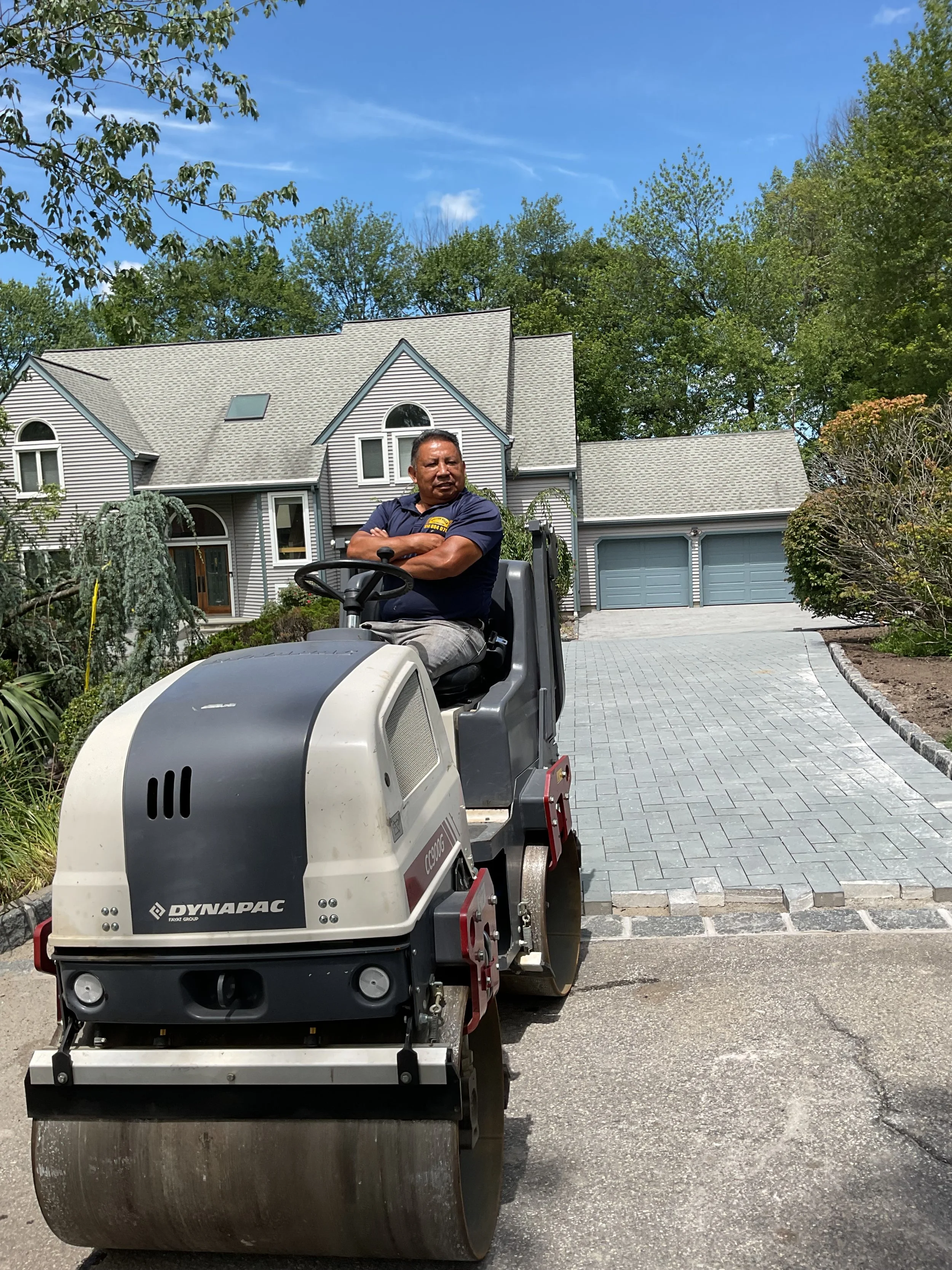A man operating a Dynapac compactor on a paved driveway in front of a large gray house with a manicured garden and trees, under a blue sky with some clouds.