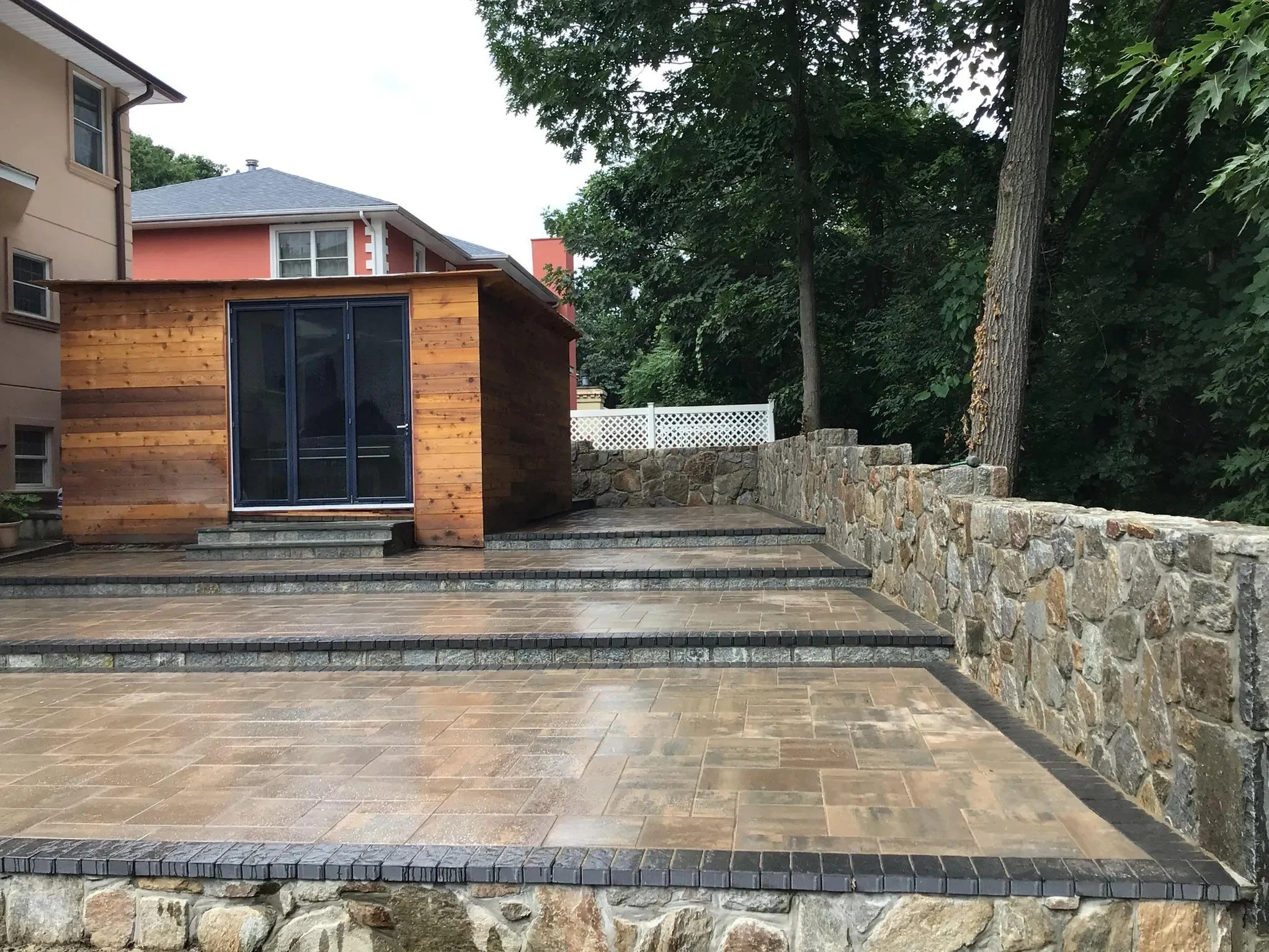 A backyard patio with stone and tile flooring, a small wooden structure with glass doors, a stone wall, and trees in the background.