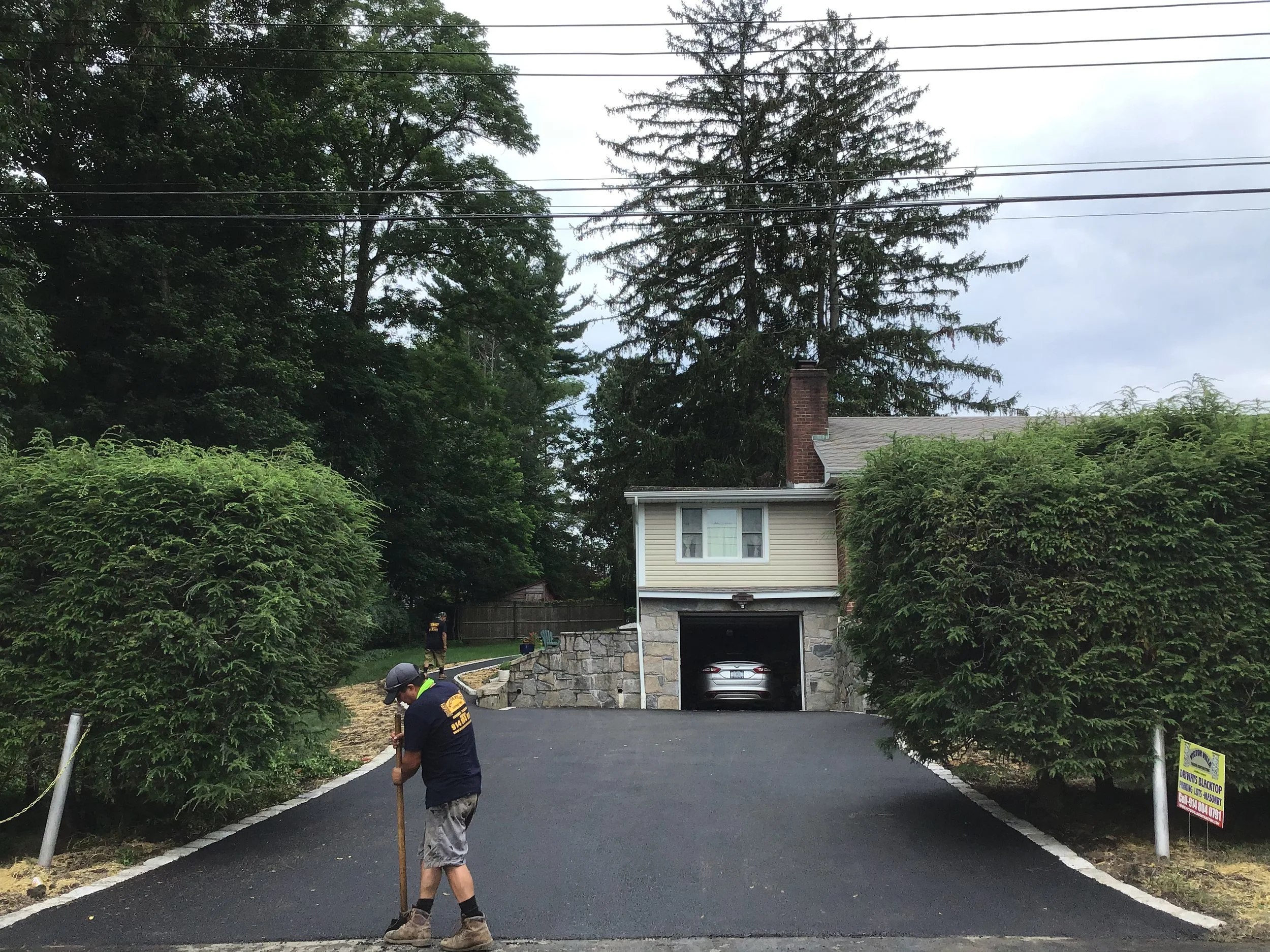 A worker is paving a driveway in front of a house, with trees and bushes surrounding the area.