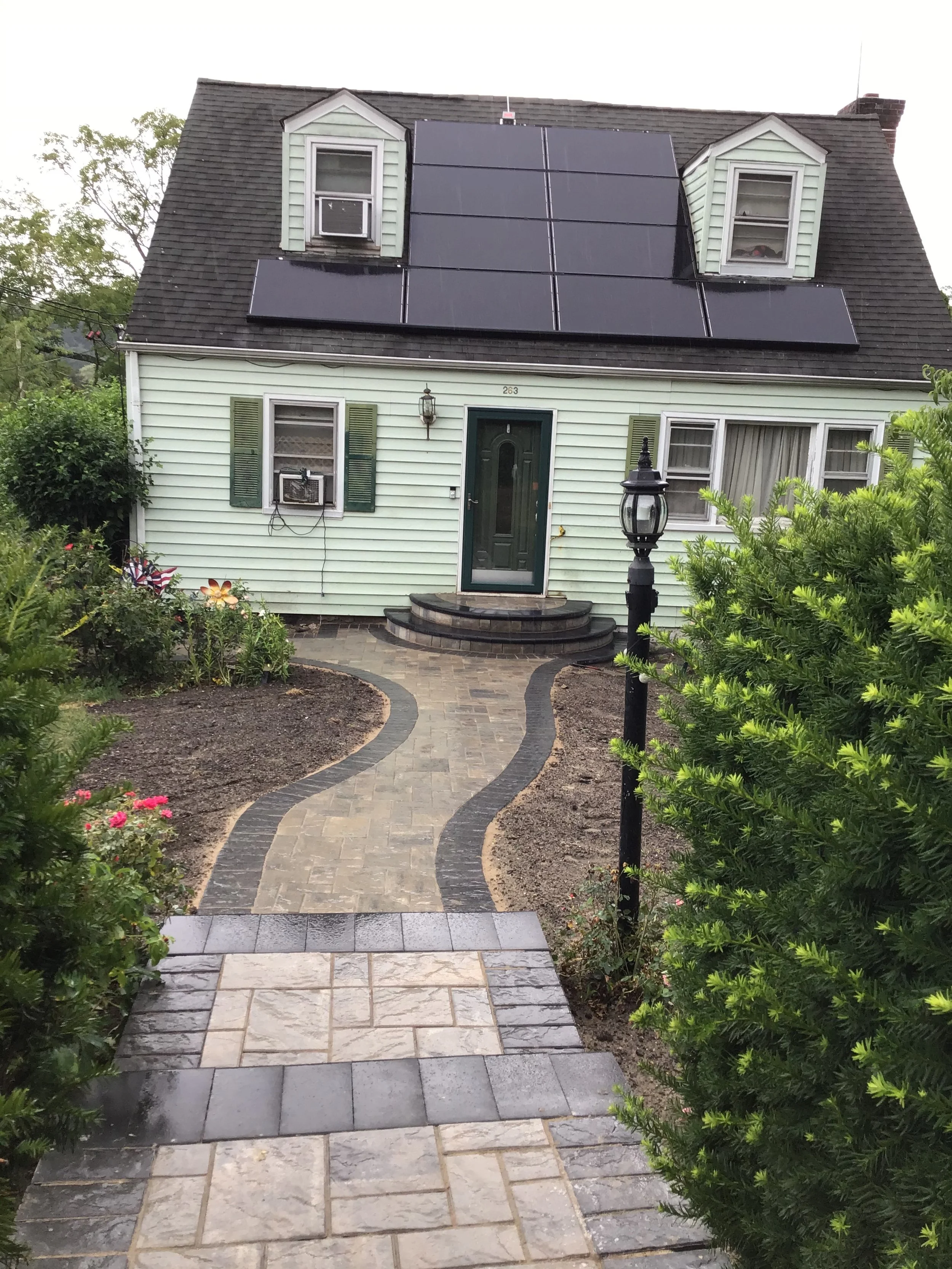 A house with light green siding and a dark green door, featuring solar panels on the roof, with a winding stone pathway leading to the front steps, surrounded by green bushes and flowers.