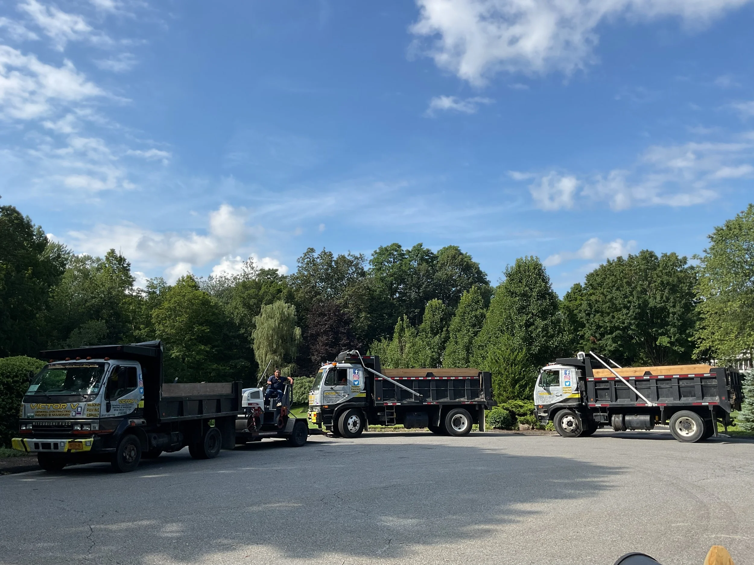 Three dump trucks parked side by side on a paved surface, with a person standing between two of the trucks in front of a green, tree-filled background under a partly cloudy sky.
