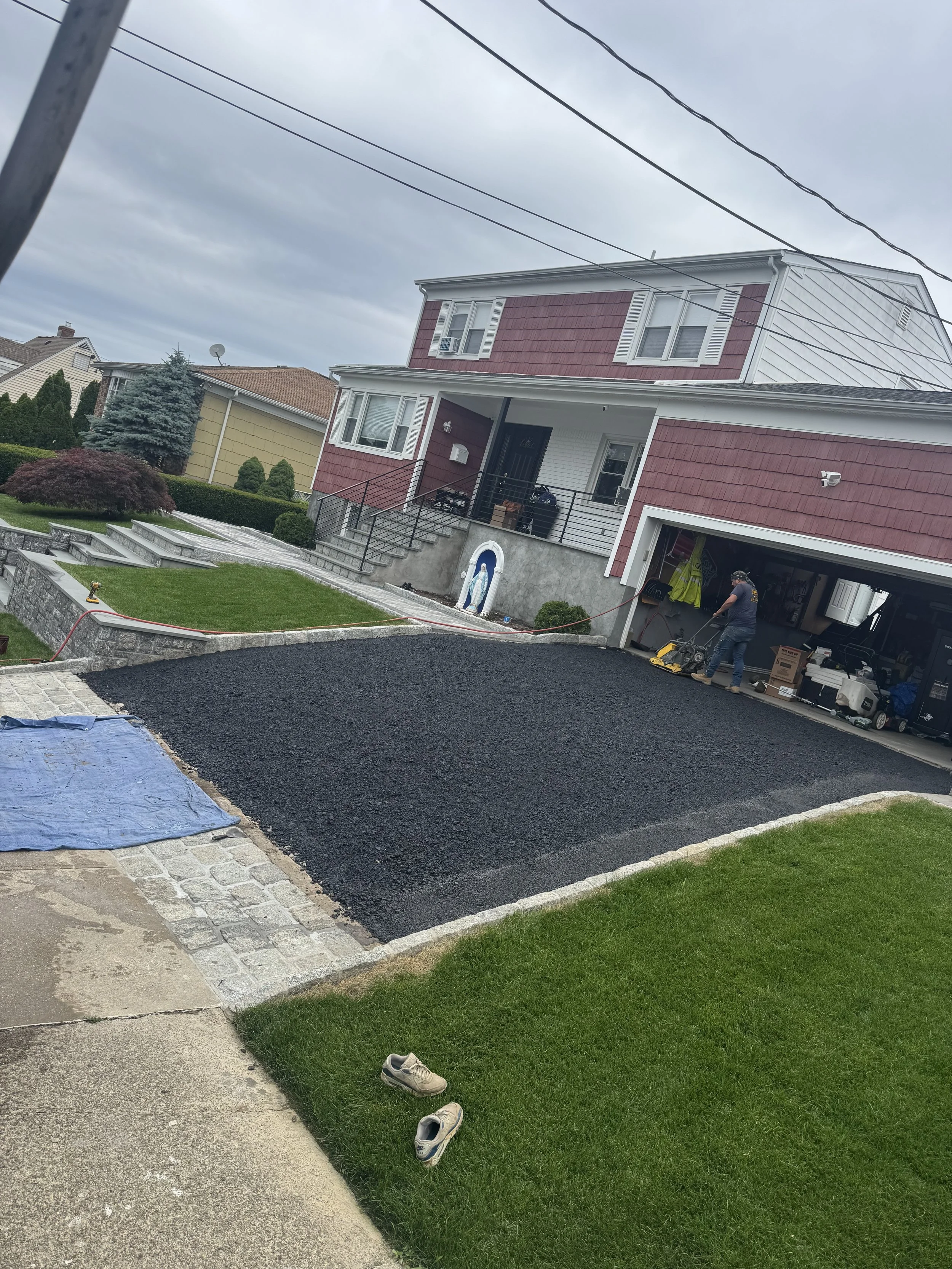 A two-story house with a freshly paved black asphalt driveway during daytime. The house has red and white siding, and there is a person working near the garage. The lawn is green, and a pair of shoes is on the grass in the foreground.