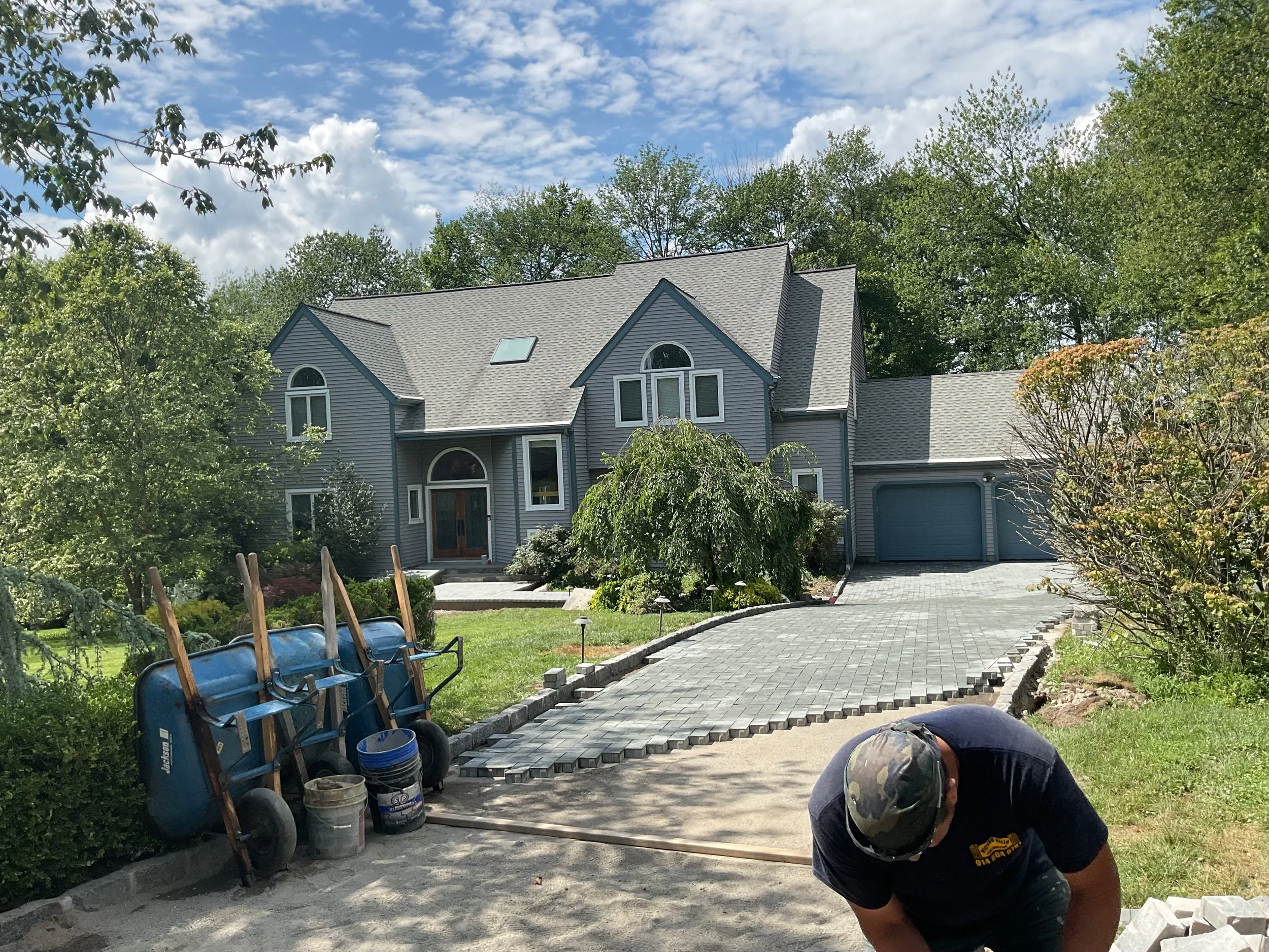 A house under construction with a worker bending over in the driveway, wheelbarrows, and construction materials in the foreground.