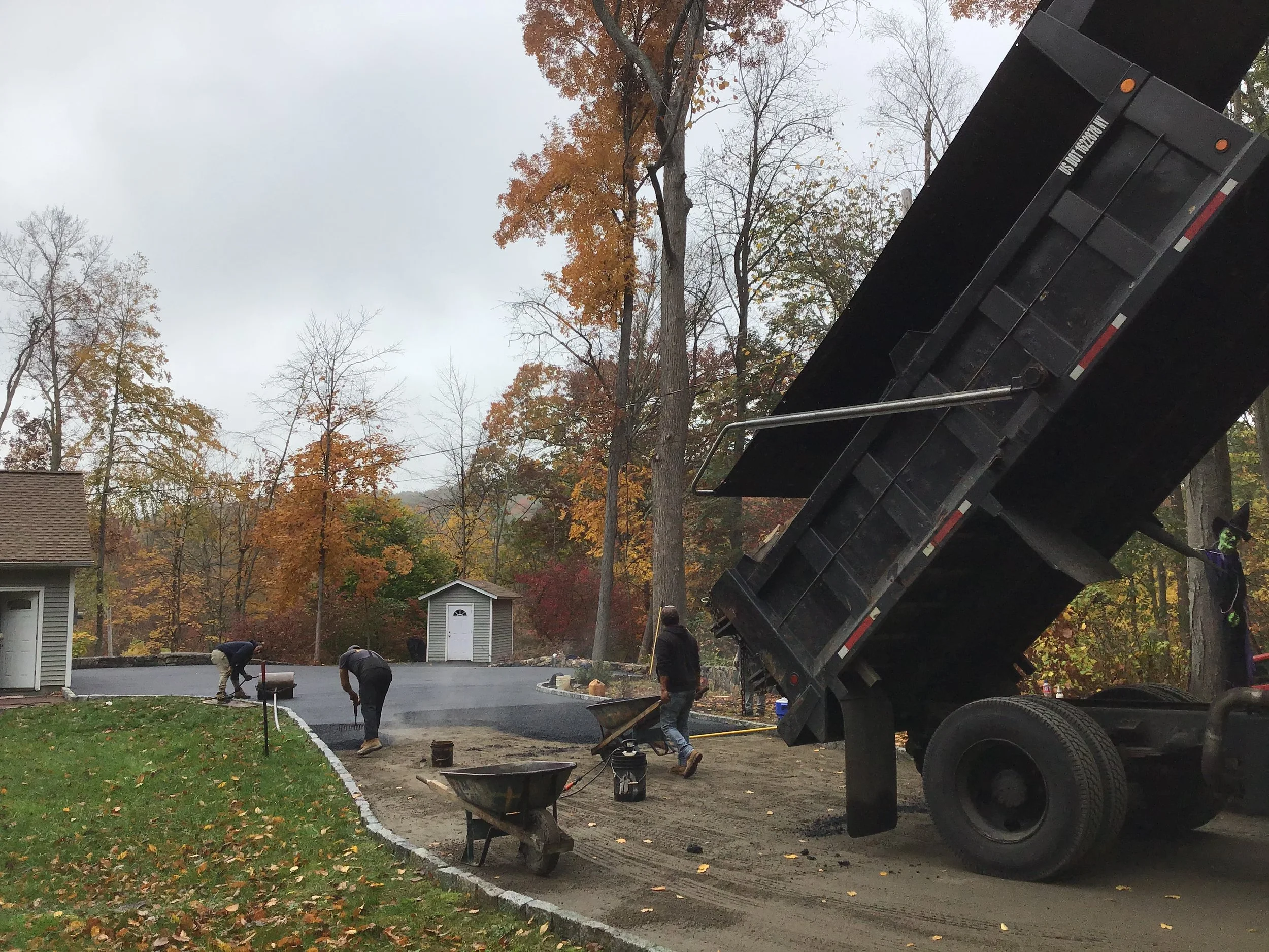 Construction workers paving a driveway with asphalt, using a dump truck to pour the asphalt, in a residential area during fall.