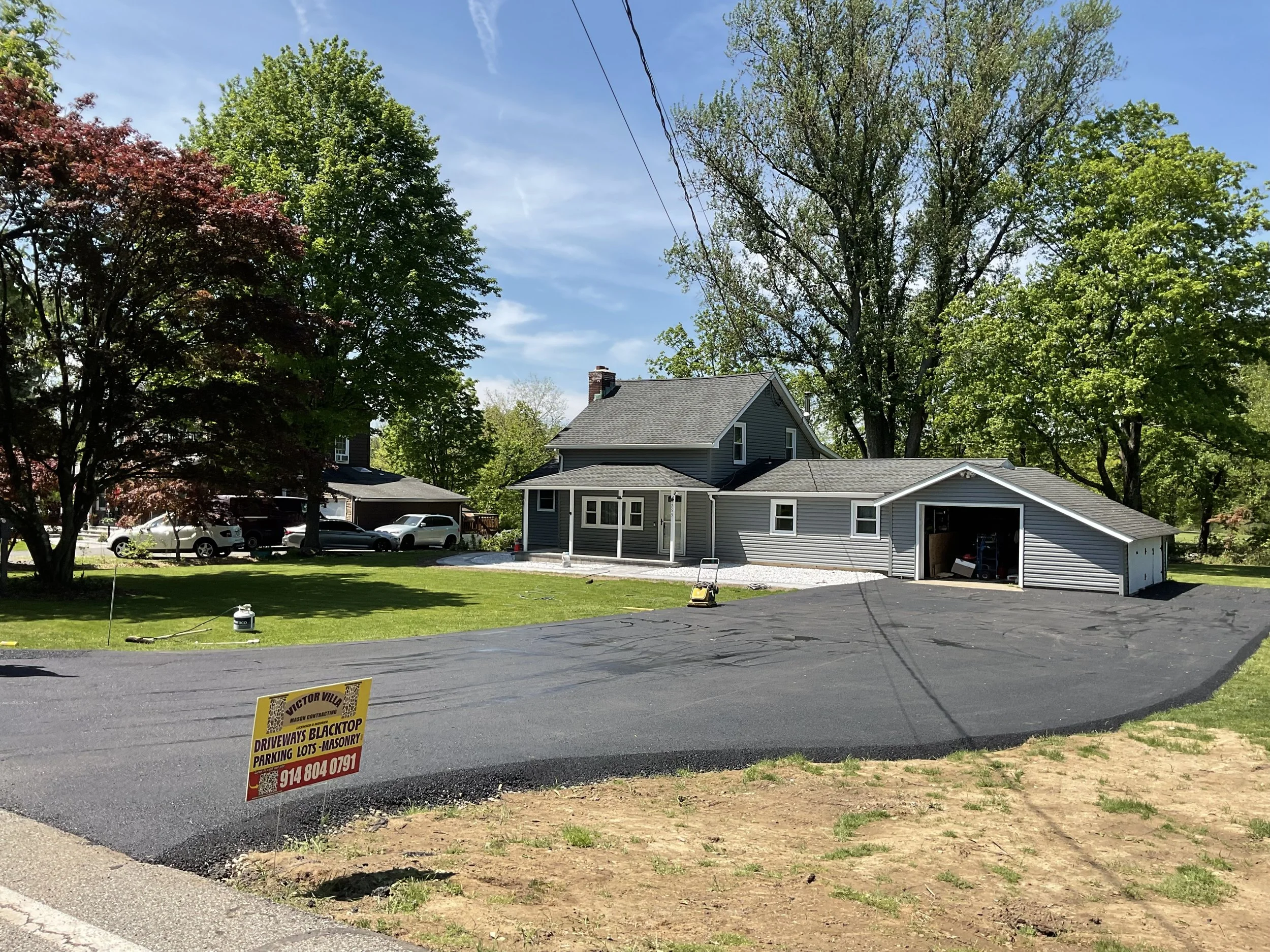 A house with a fresh asphalt driveway under construction, surrounded by green trees and a lawn, with a sign indicating paving services.