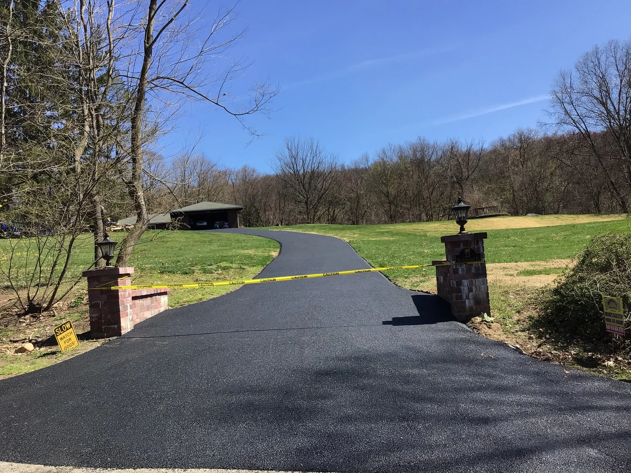 Newly paved black asphalt driveway blocked by yellow caution tape, with brick pillars and lamp posts on either side, leading to a garage surrounded by a grassy yard with trees and hills in the background.
