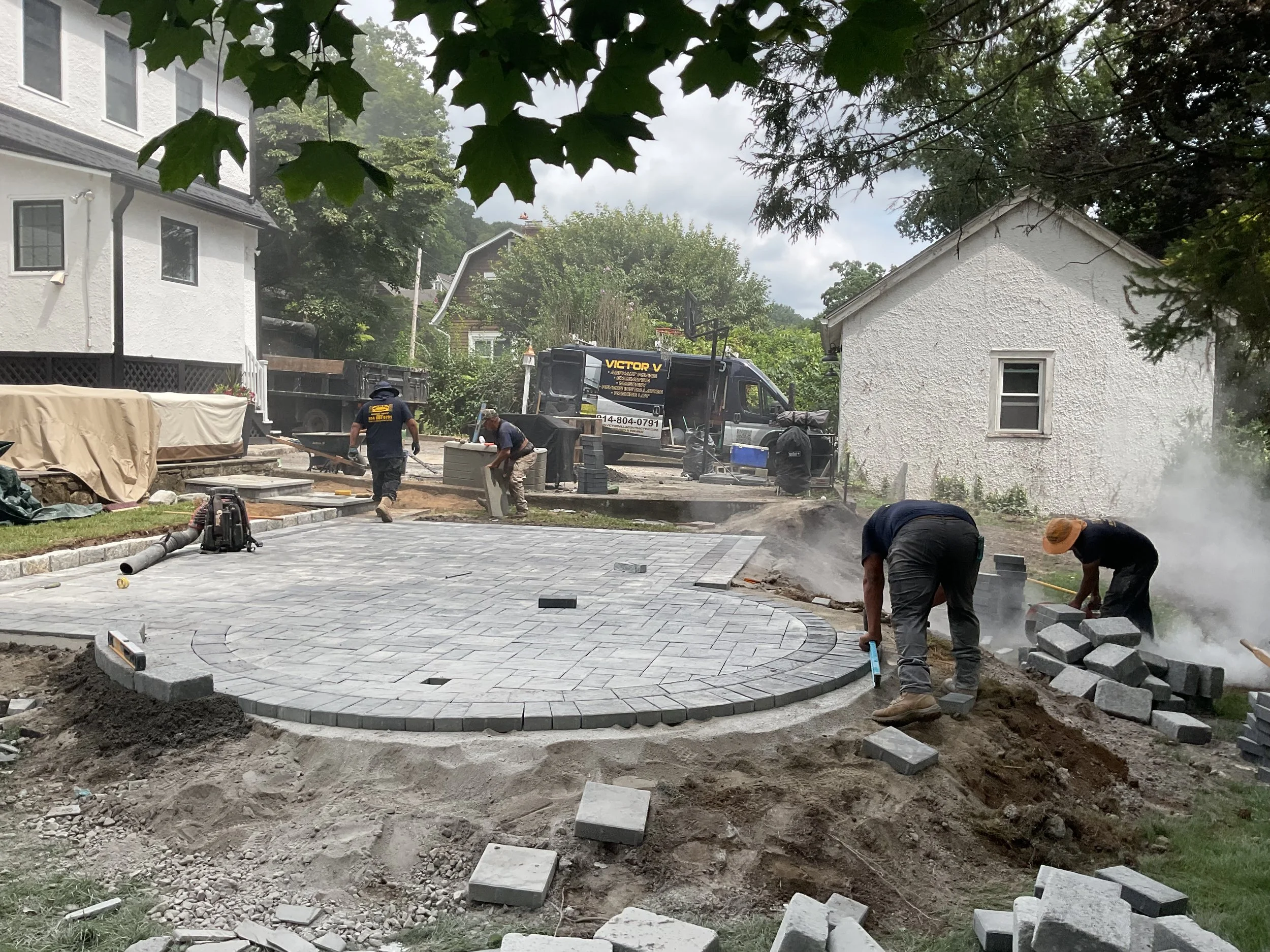 Workers laying gray stone pavers to create a curved patio area in a backyard under a cloudy sky, with trees and houses in the background.