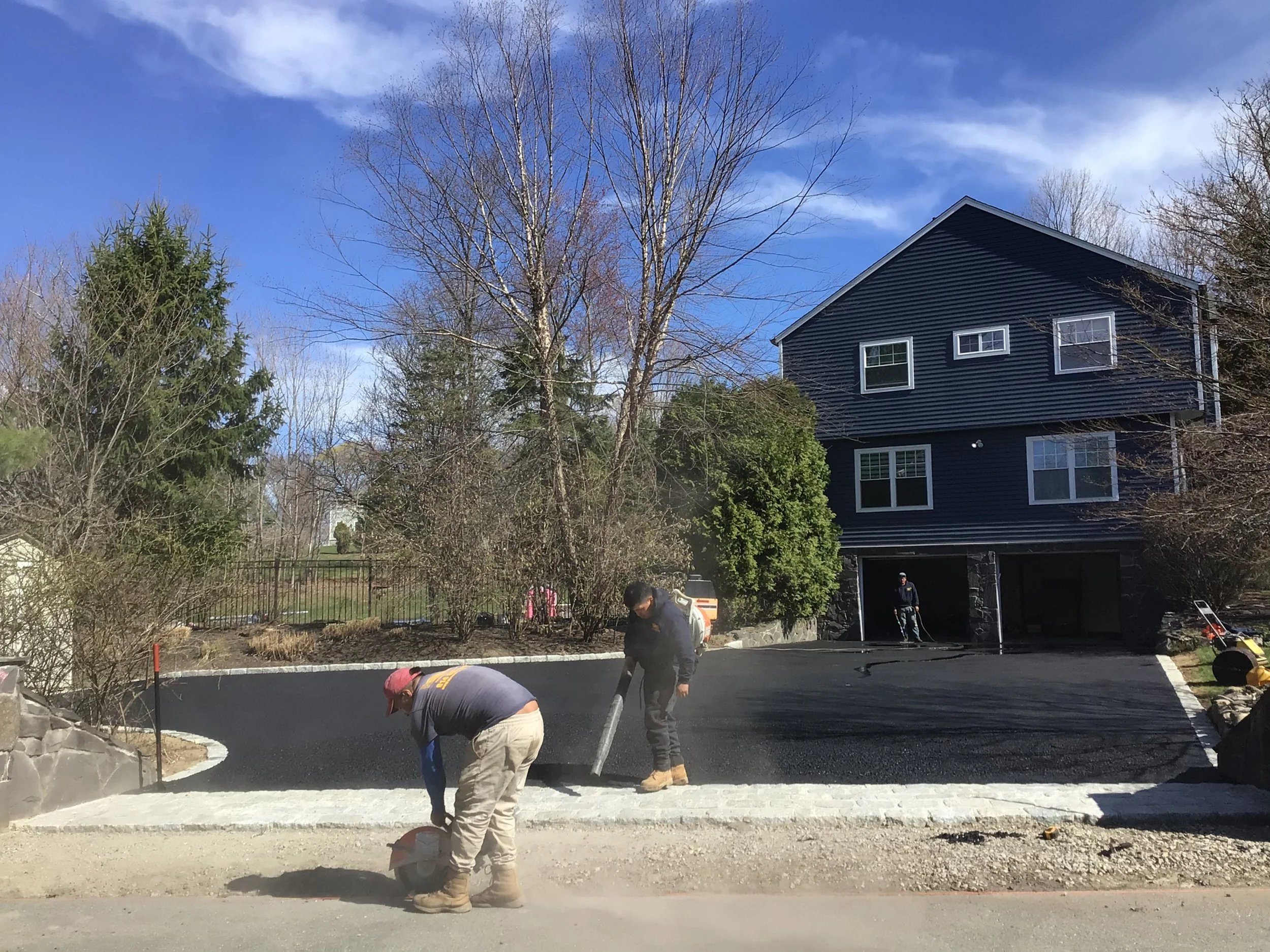 Workers paving a driveway in front of a blue house with trees and a partly cloudy sky in the background.
