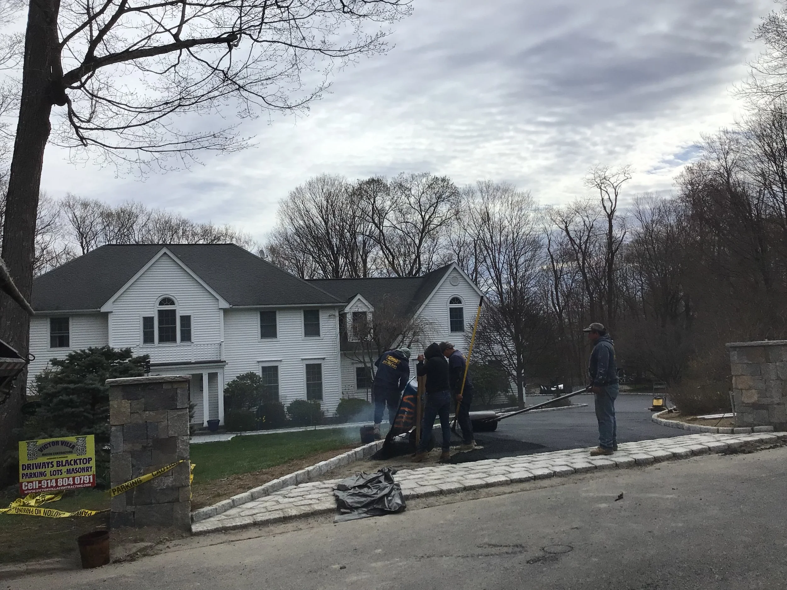 Group of workers laying cobblestone on the driveway outside a white house with black shutters, surrounded by leafless trees.