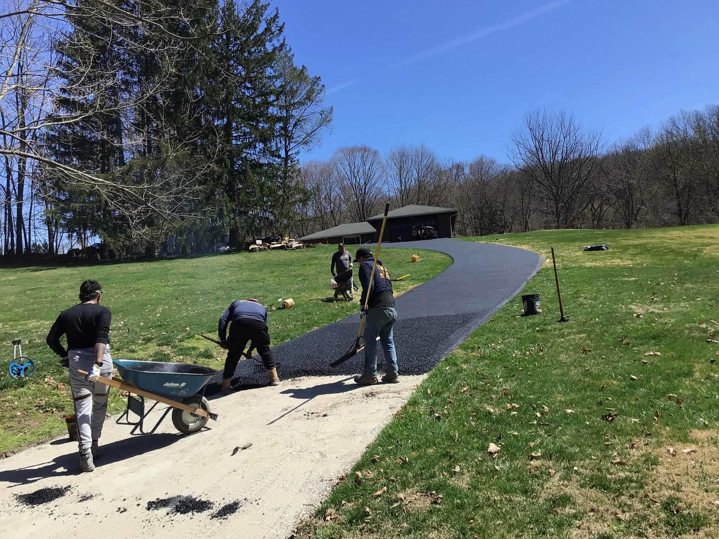 Workers laying asphalt on a winding pathway in a park or yard, with trees and a small building in the background under a clear blue sky.