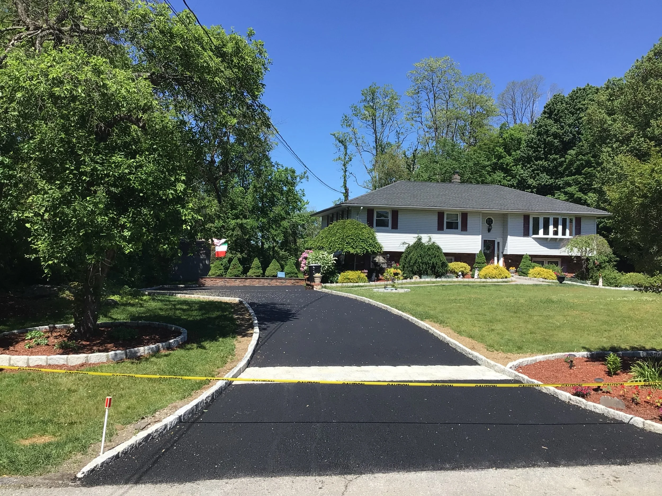 A newly paved black asphalt driveway curves toward a house with white siding, surrounded by green lawn, trees, and shrubs under a clear blue sky.
