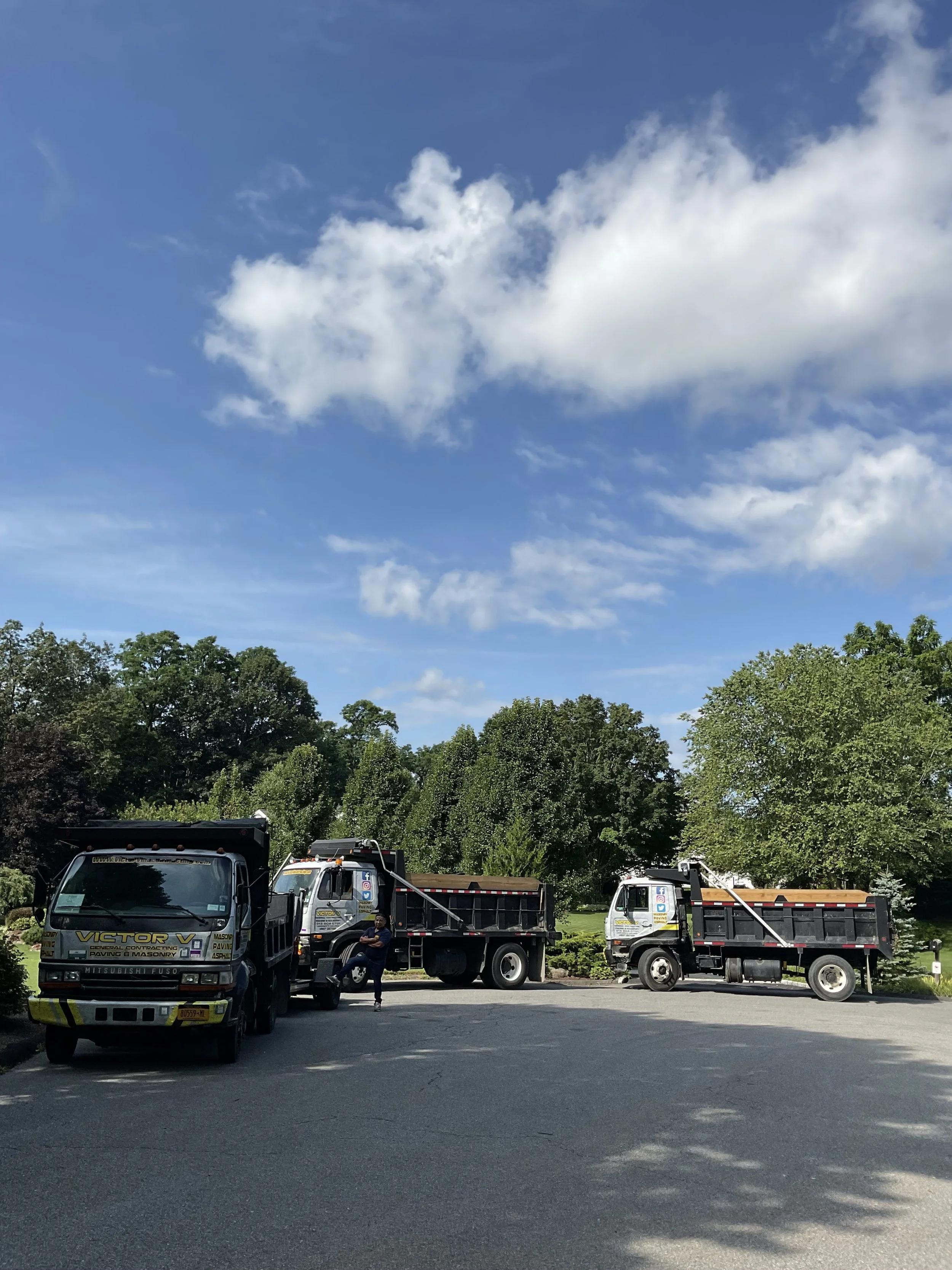 Three trucks parked on a paved area under a partly cloudy blue sky, with trees in the background. A man is standing near the front of the first truck.