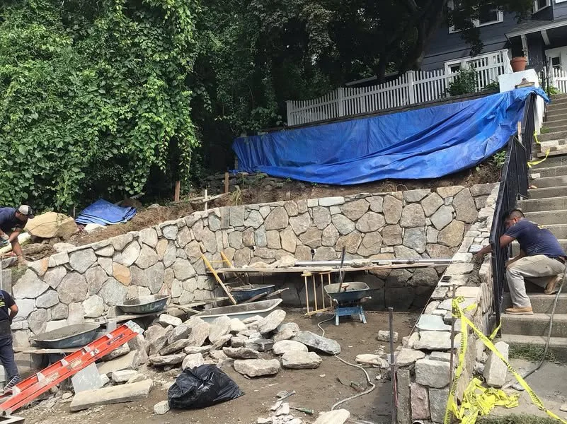 Construction workers building a retaining wall with large stones in a backyard, with a blue tarp covering part of the slope and stairs on the right side.