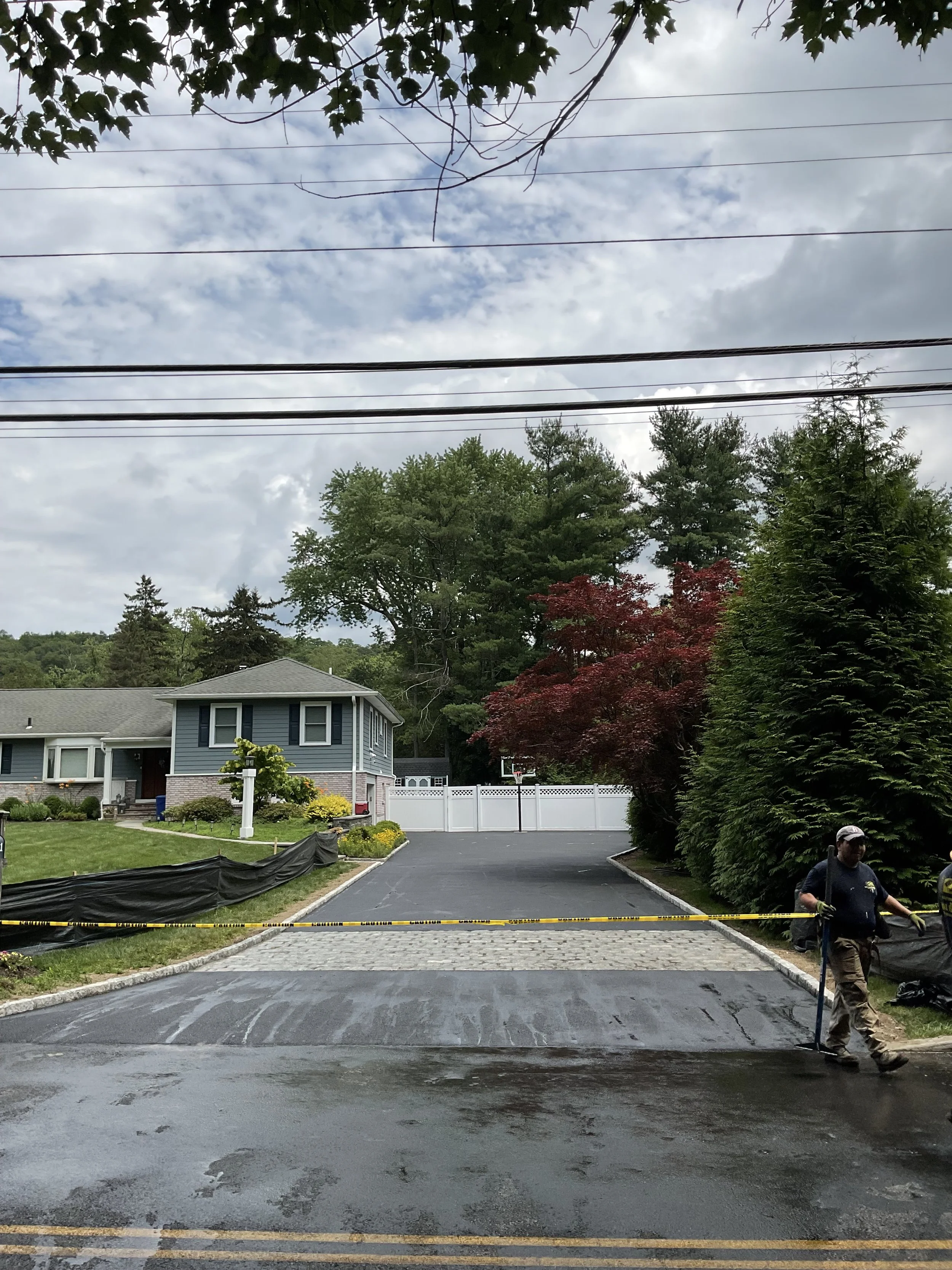 A residential area with a newly paved driveway, police tape surrounding it, a worker standing near the tape, green trees, a house in the background, and a partly cloudy sky.