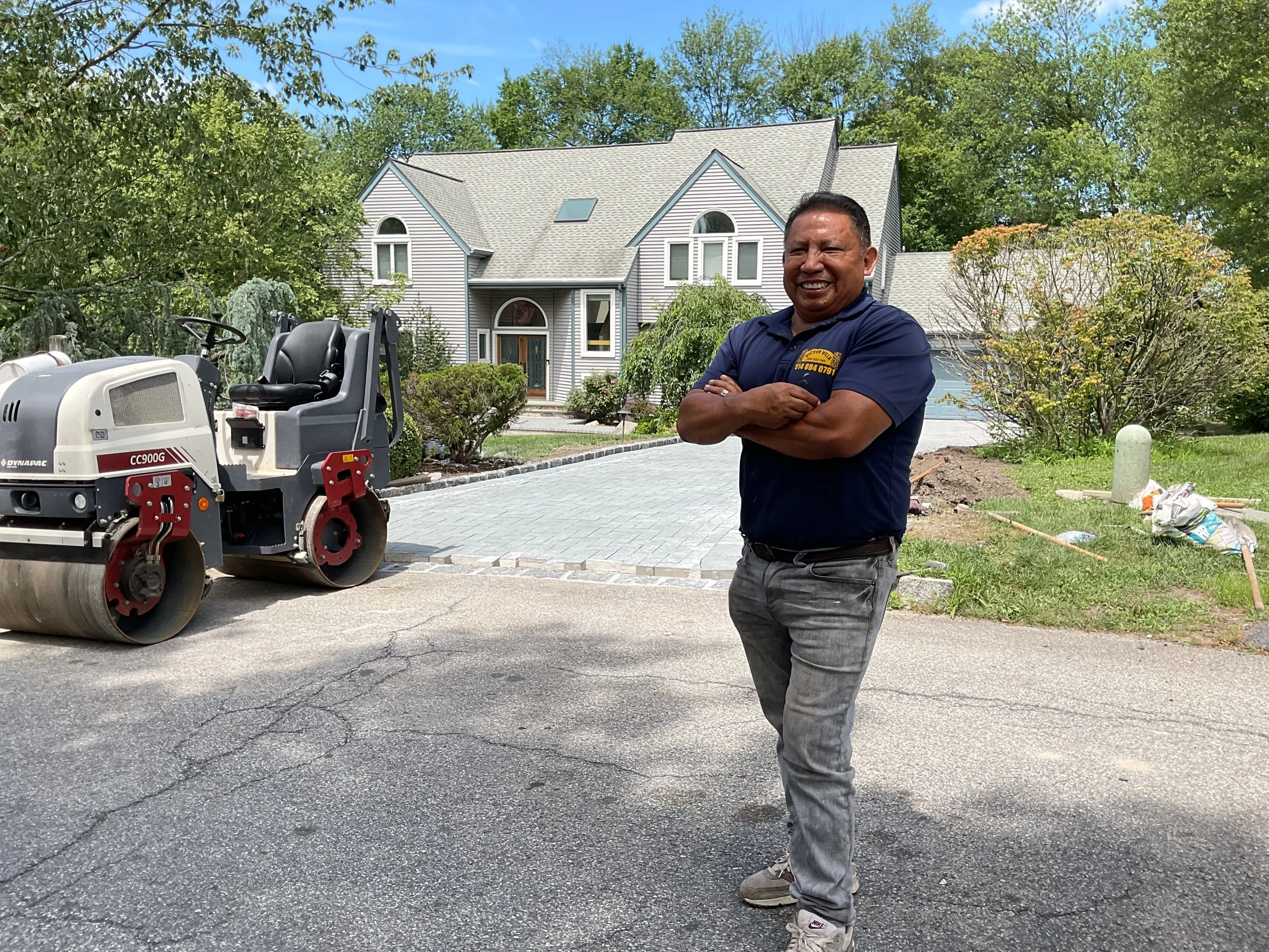 A smiling man stands with arms crossed in front of a driveway with construction equipment, in a residential neighborhood with a large house and trees in the background.