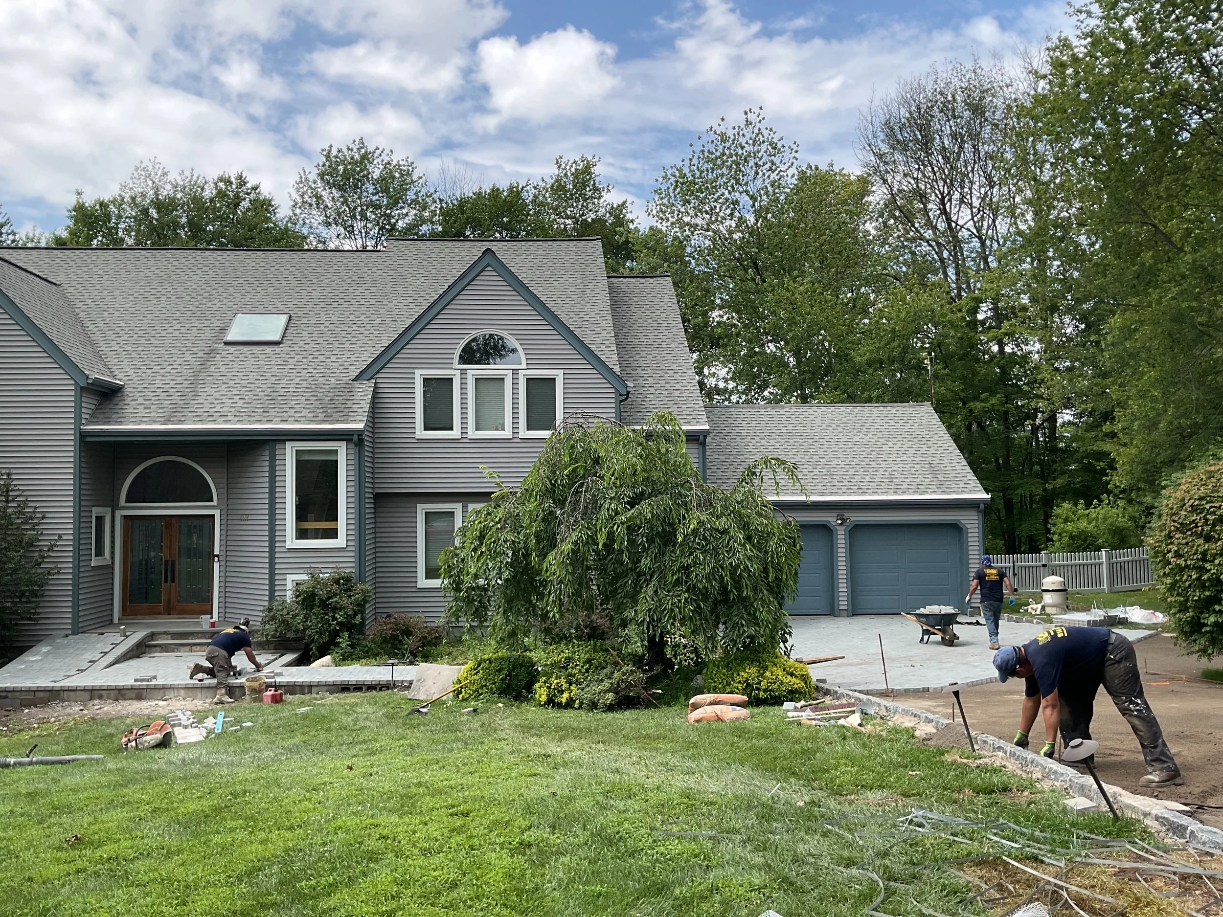 Construction workers paving driveway in front of a blue house with a landscaped yard and trees.