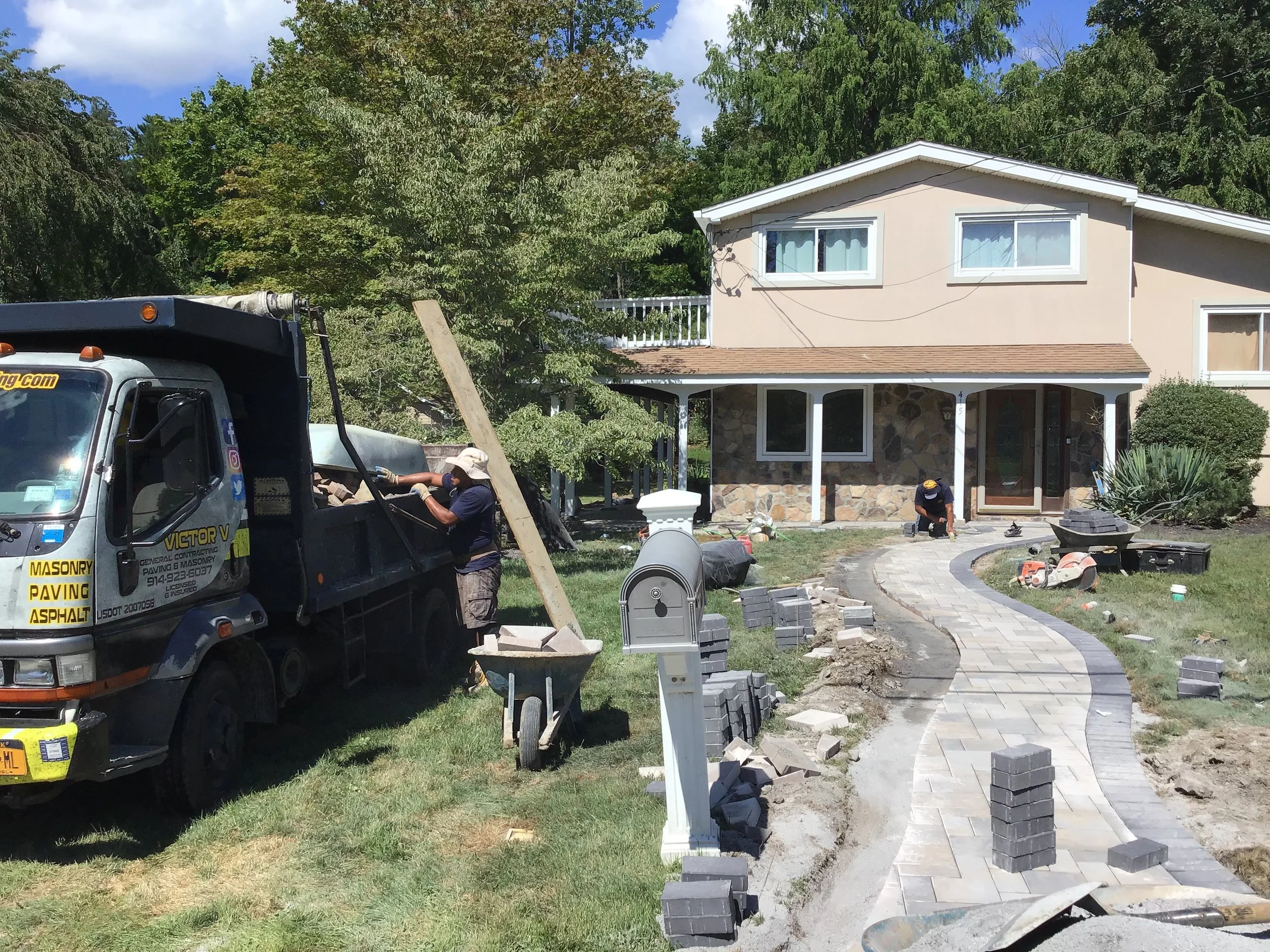 Construction workers building a curved stone pathway in front of a beige house with stone accents, surrounded by green trees and a blue sky.