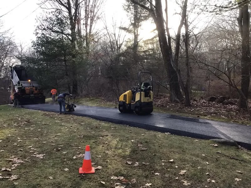 Workers in reflective vests paving a road in a park-like setting with trees, a traffic cone, and construction equipment including a roller compactor.