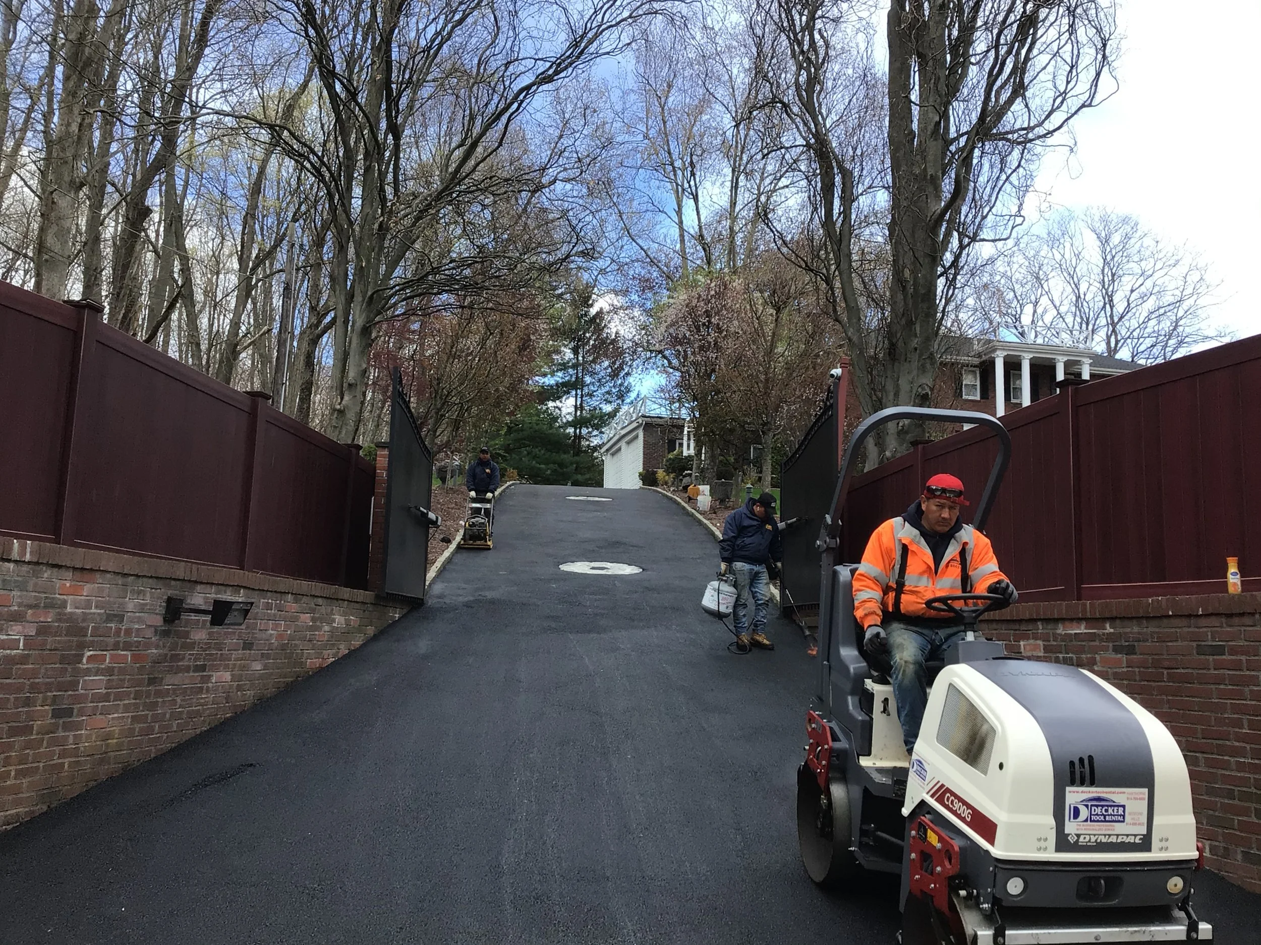 Workers paving a long, inclined driveway with asphalt, flanked by brick and wooden fences, in a suburban neighborhood with trees and houses in the background.