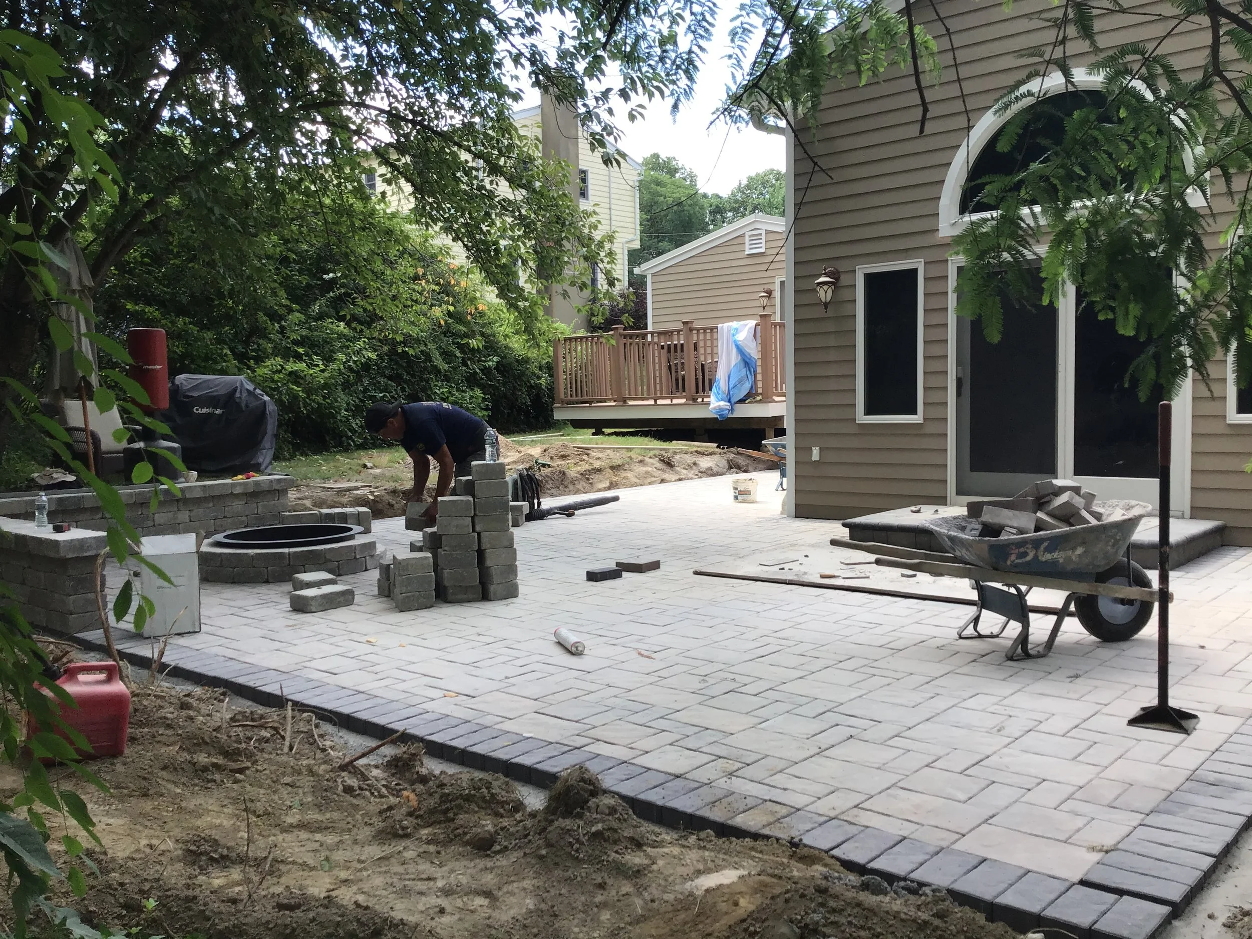 A man installing paver bricks on a backyard patio, with a wheelbarrow and stack of bricks nearby, during daytime.