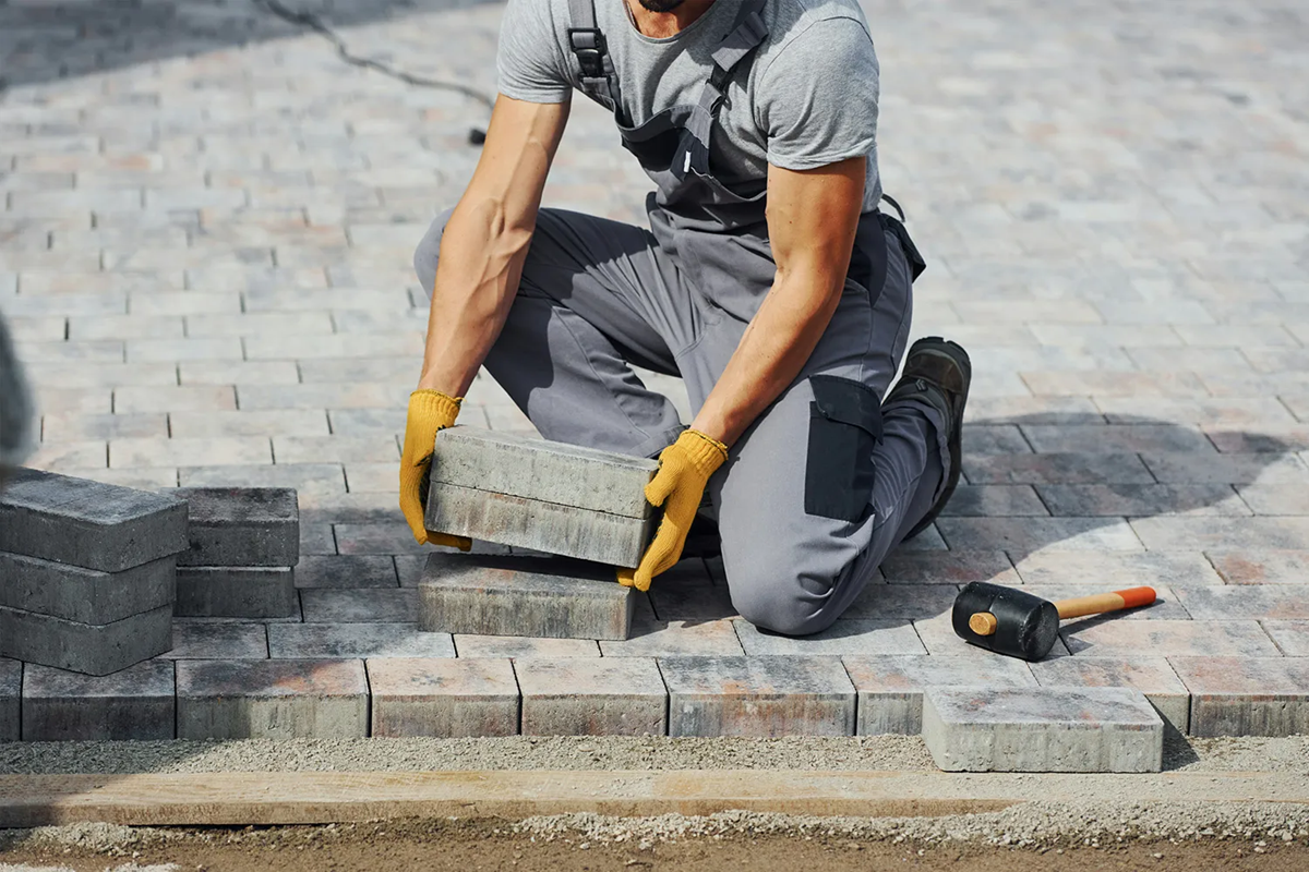 Worker installing bricks on a paved surface, kneeling with yellow gloves, using a rubber mallet and stacking bricks.