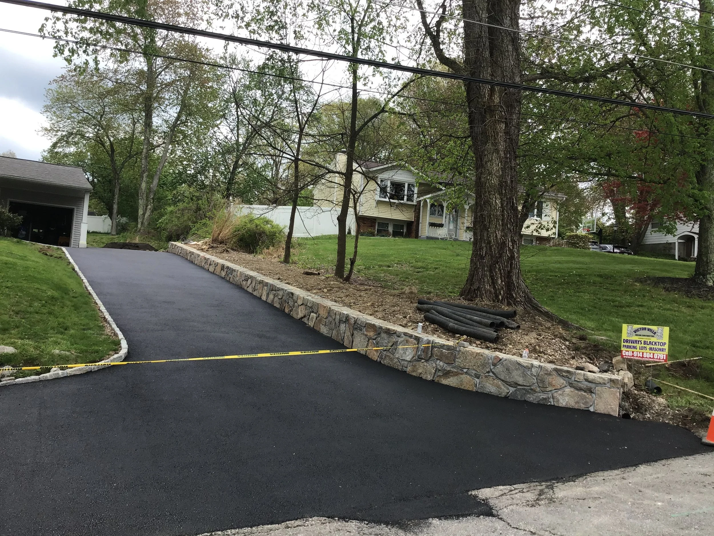 Freshly paved driveway with a stone retaining wall on the right side, in front of a house with trees and a yard, with caution tape and a construction sign nearby.